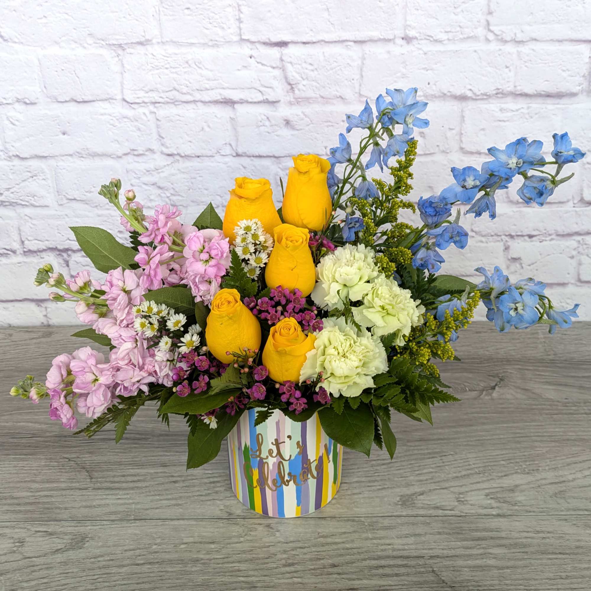 Colorful bouquet of yellow roses, pink stock, and blue delphinium in a striped container