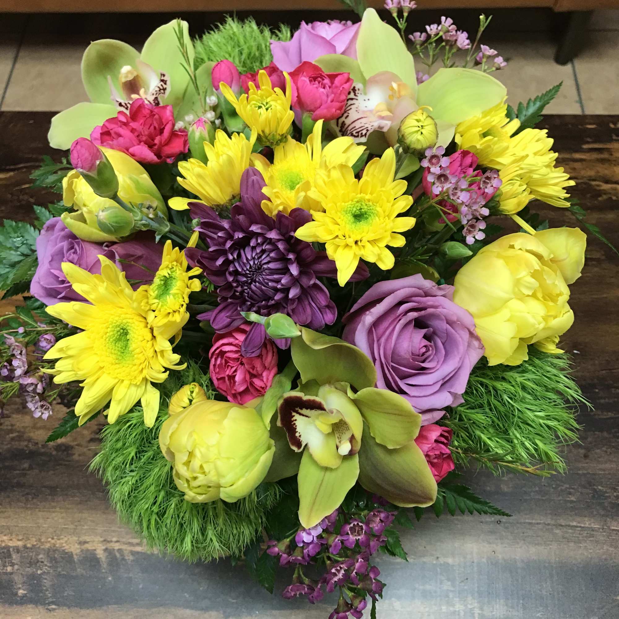 Compact arrangement of yellow daisies, green orchids, lavender roses, and pink flowers in a round design.