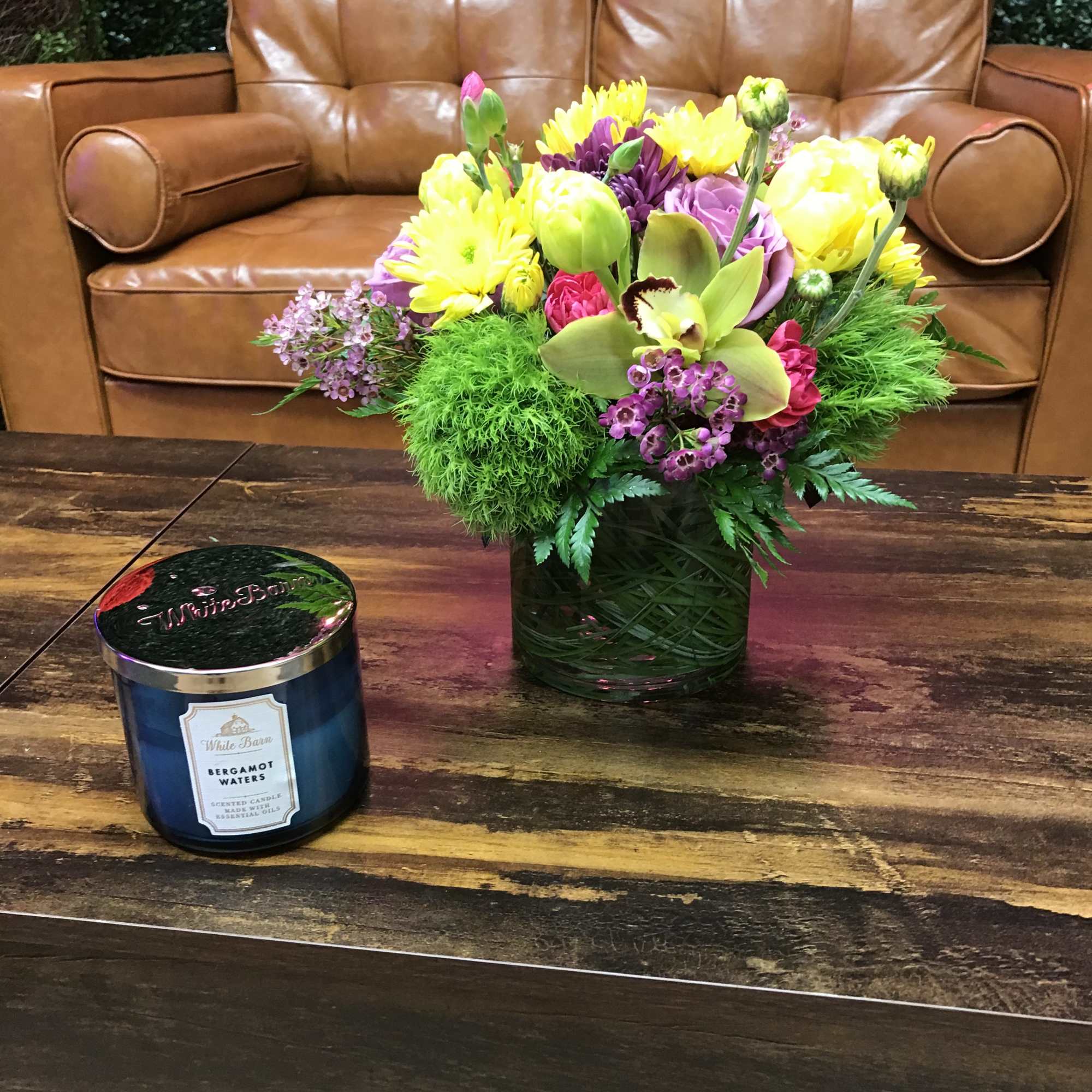 Bright mixed flower arrangement in a glass vase next to a blue candle on a wooden table