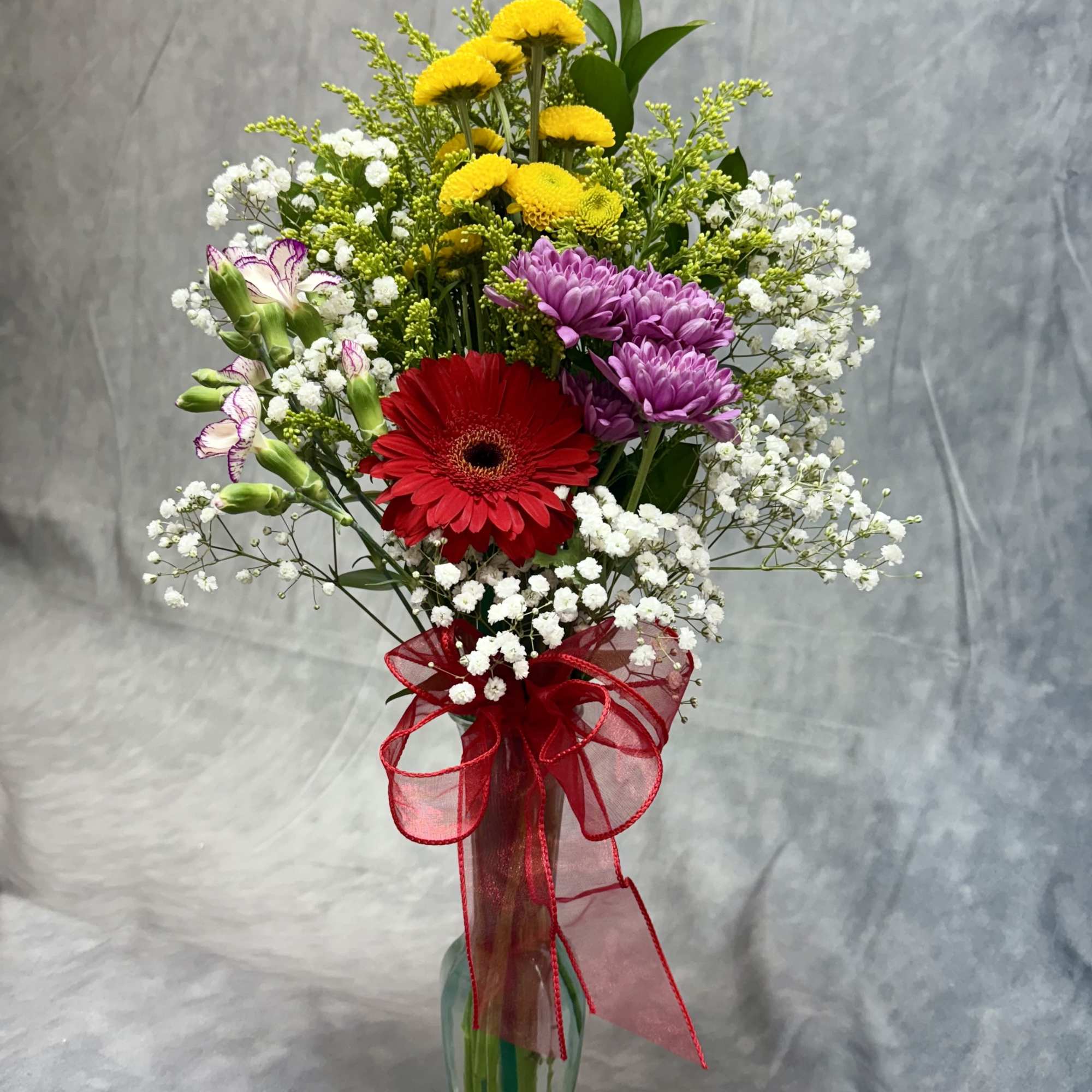 Mixed bouquet of red, yellow, and purple flowers in a glass vase with a red ribbon