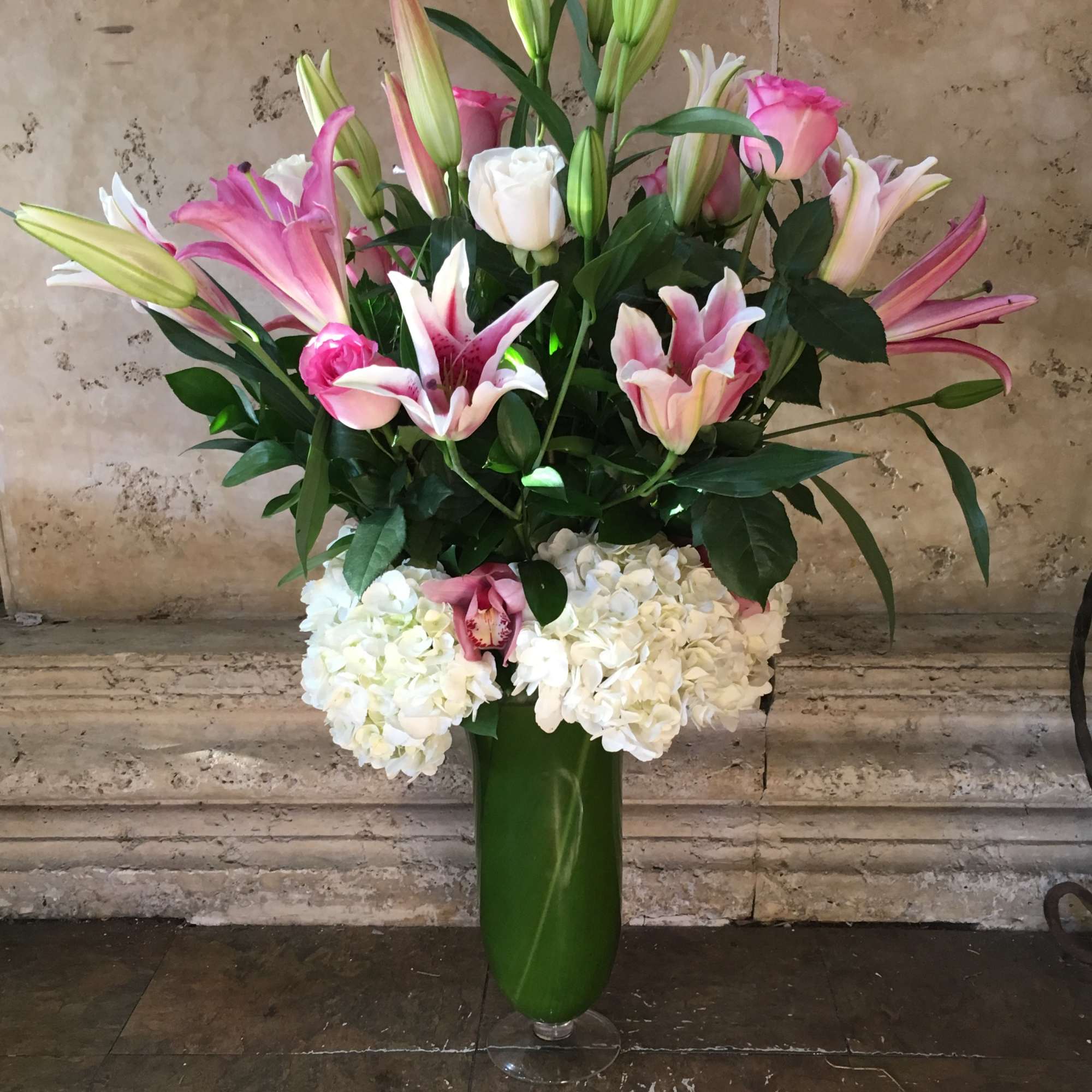 Pink lilies and white roses in a tall green vase with white hydrangeas below