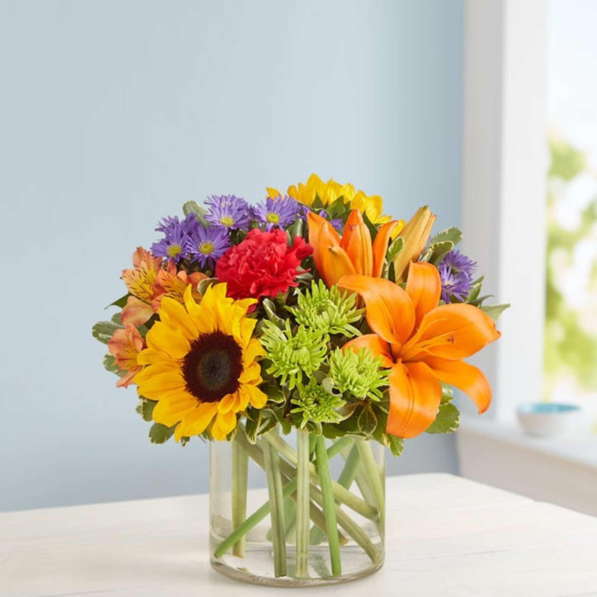 Mixed bouquet with sunflowers, lilies, and purple daisies in a glass vase
