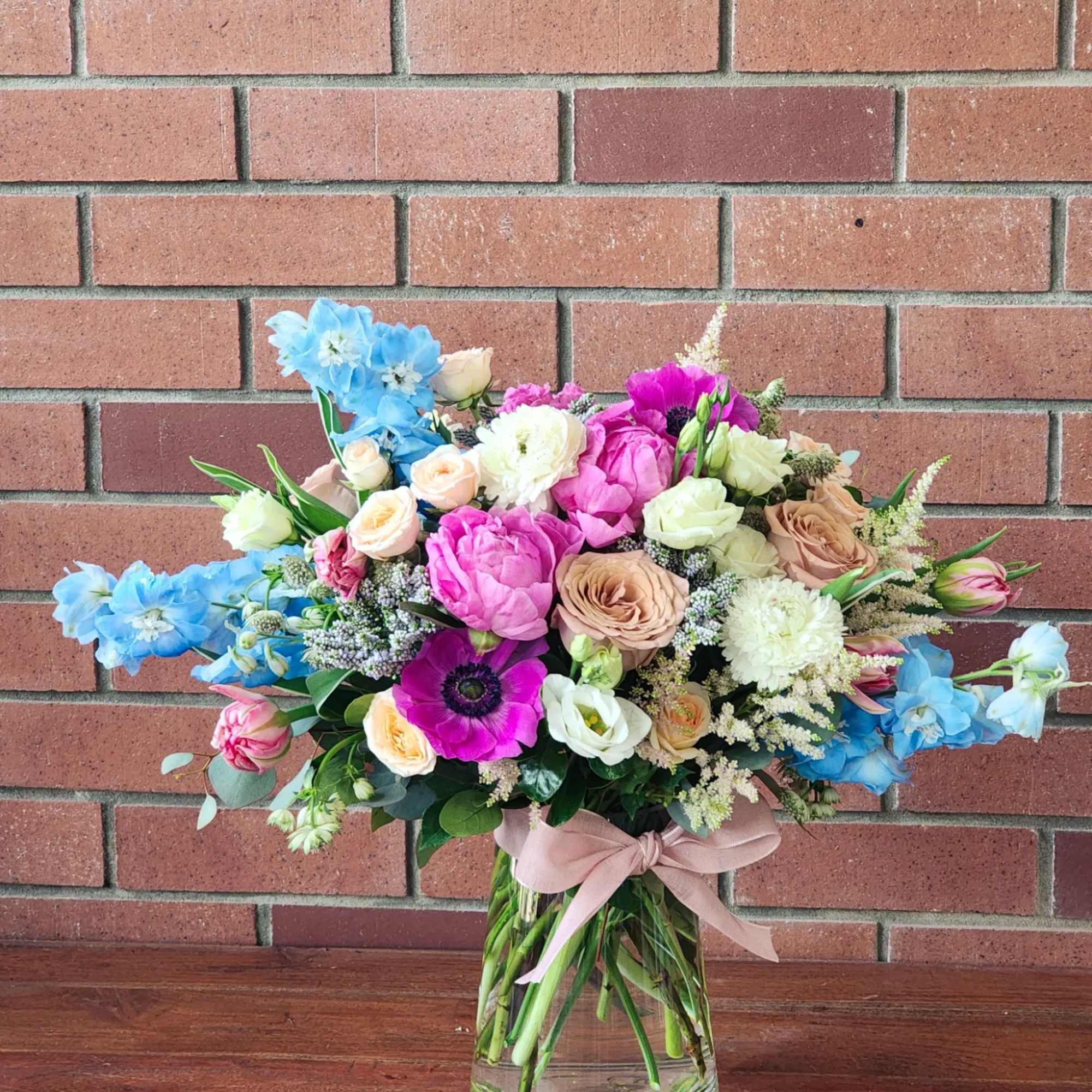 Colorful mixed bouquet of pink, peach, white, and blue flowers in a clear glass vase with a bow