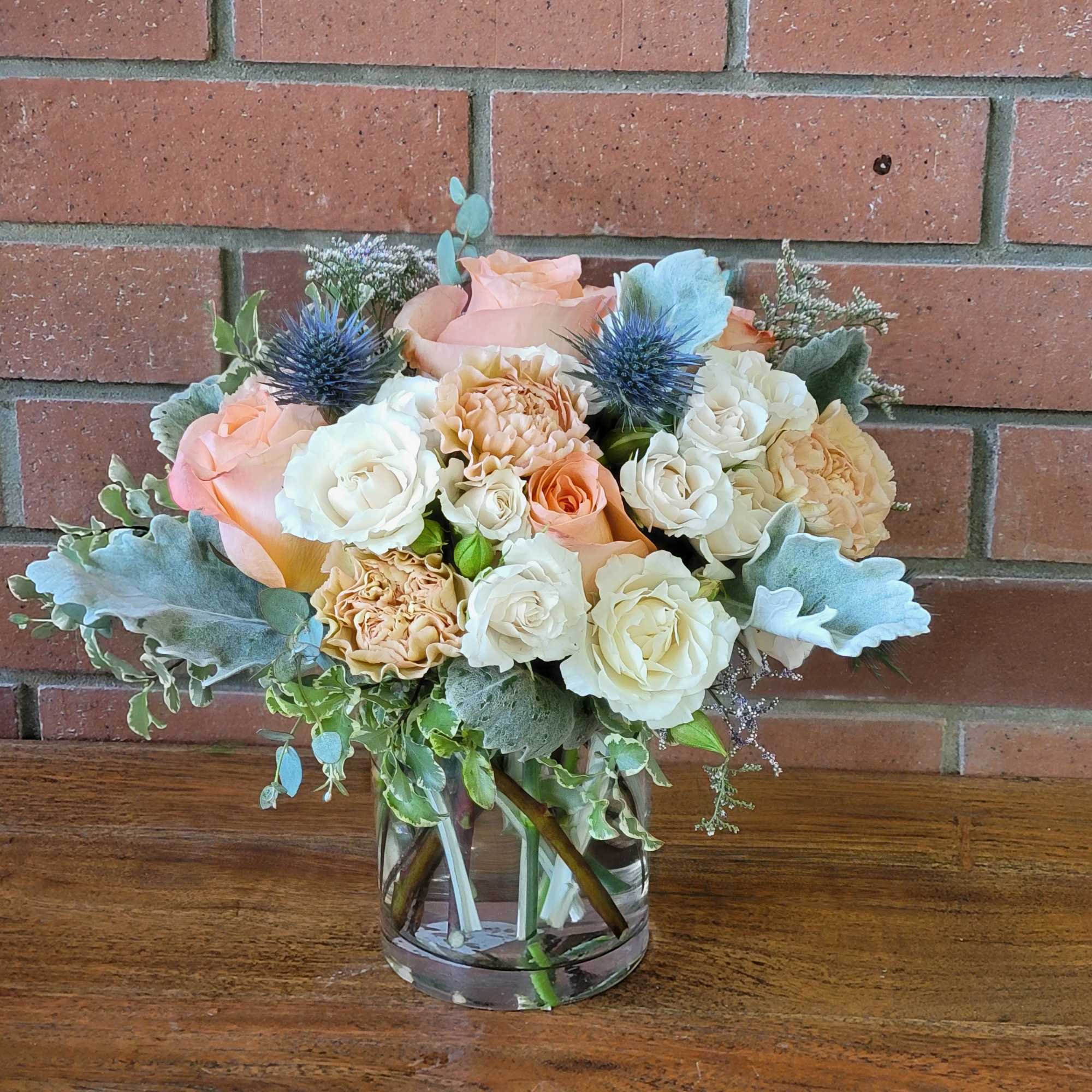 Peach and cream rose arrangement with blue thistles in a clear glass vase