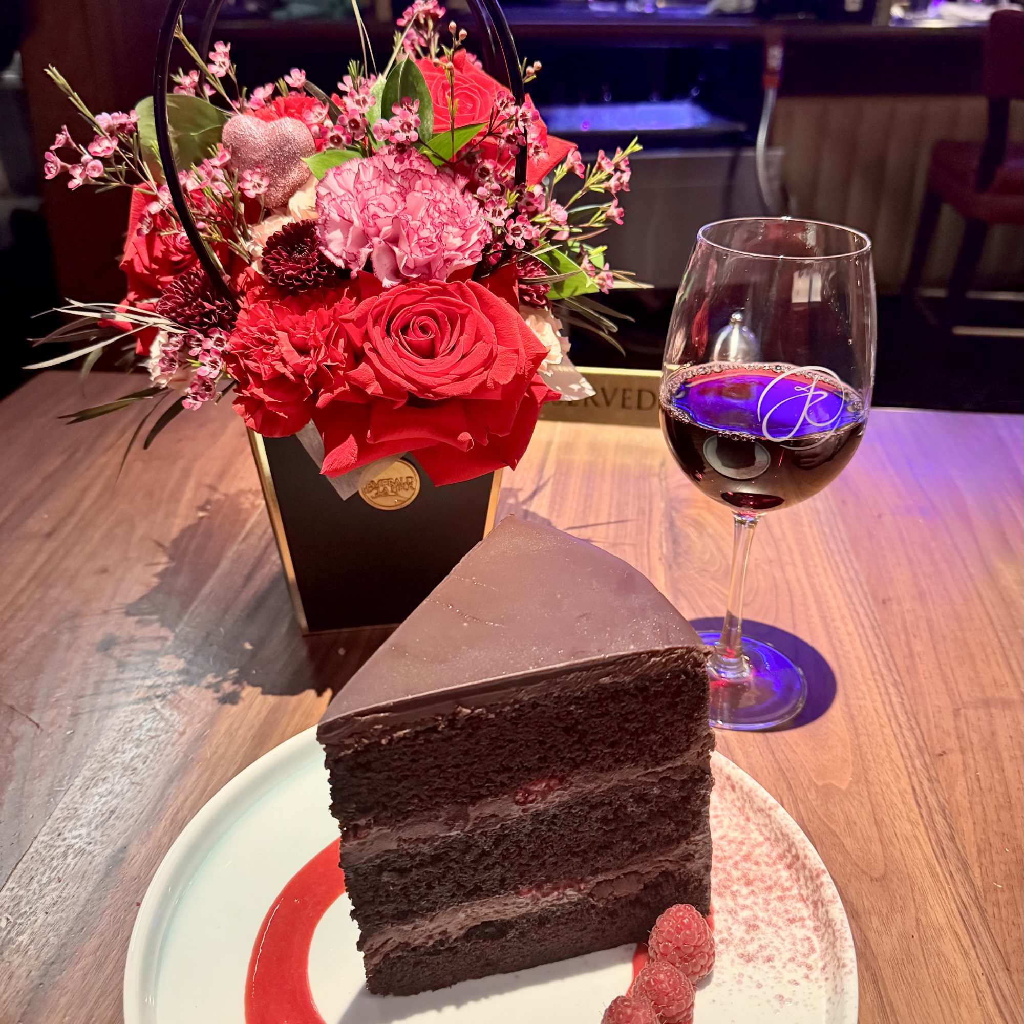 Red and pink flower arrangement beside a wine glass and chocolate cake slice