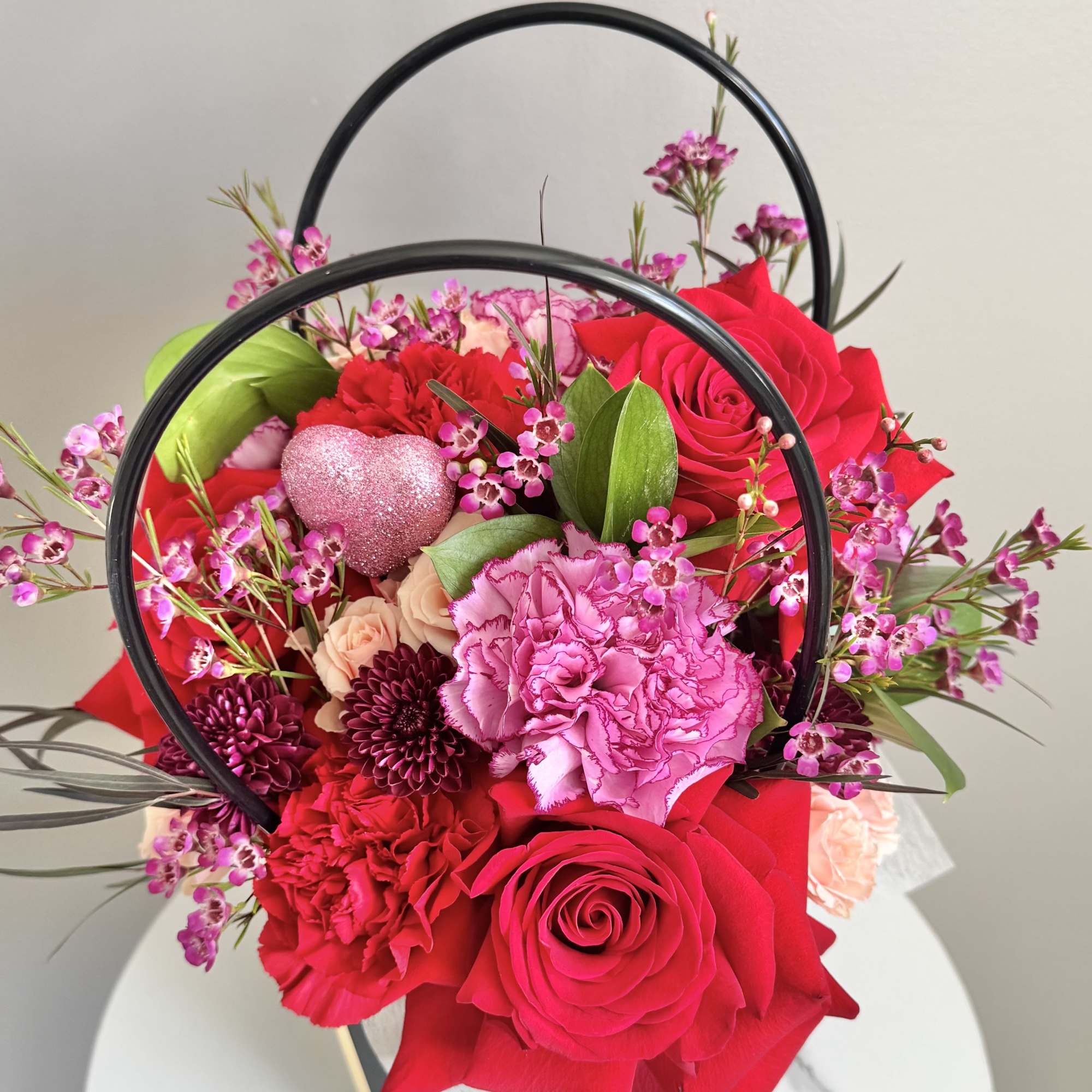 Pink and red floral arrangement in a black handled container with a glitter heart