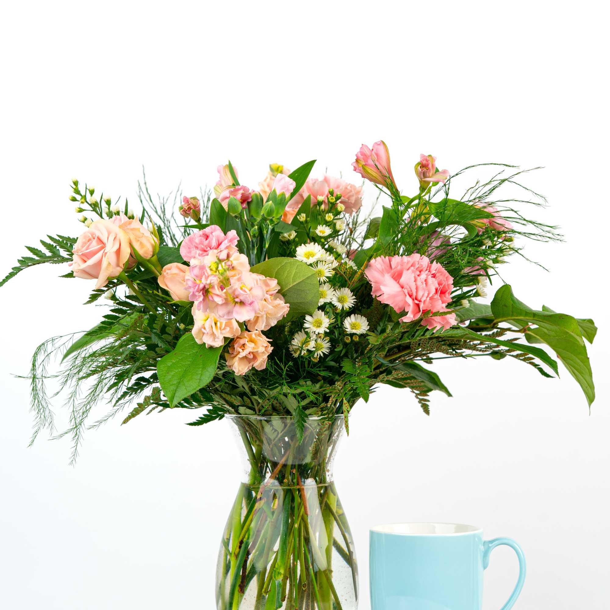 Mixed bouquet of pink and peach flowers in a clear vase beside a light blue mug