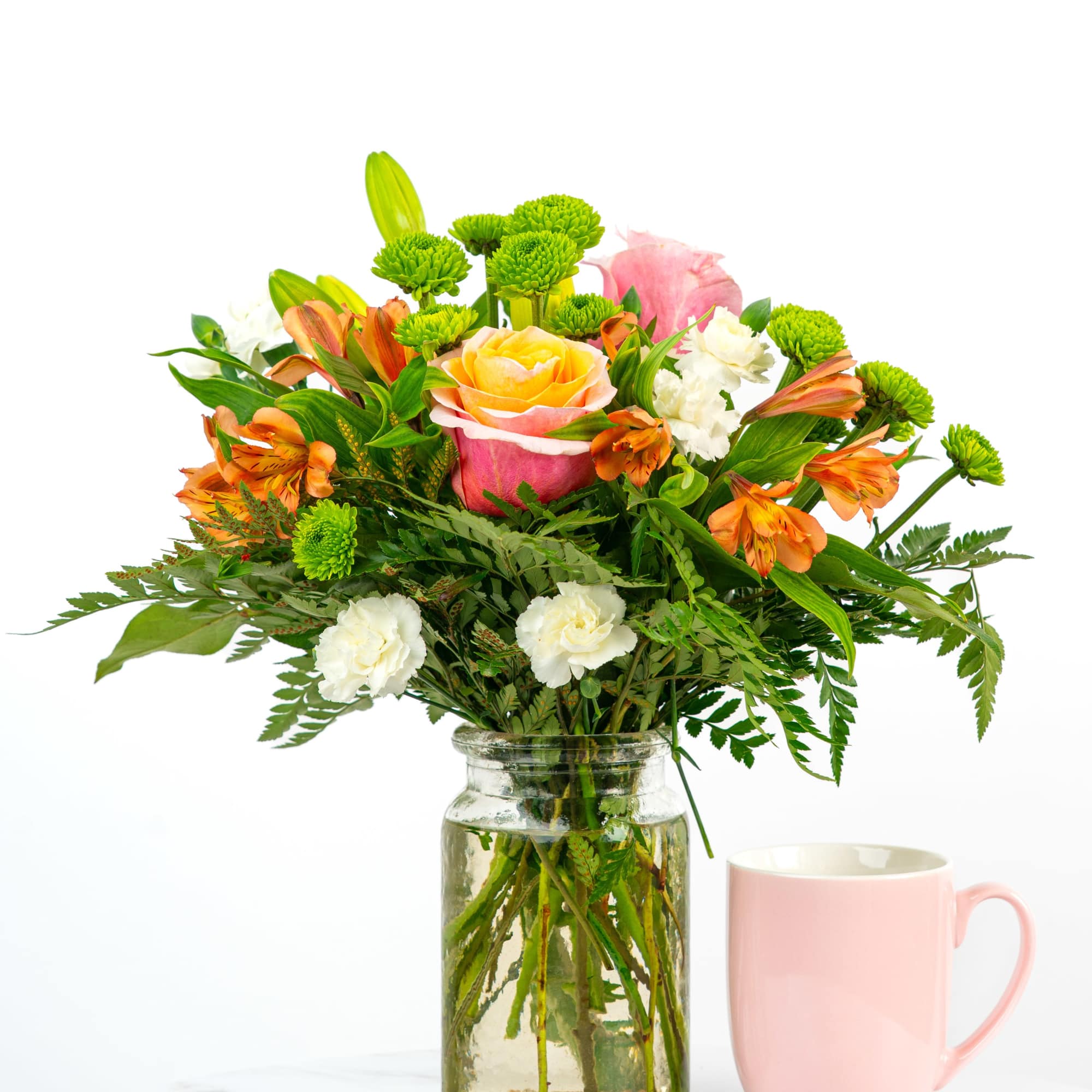 Mixed bouquet of roses, lilies, and green button mums in a glass jar beside a pink mug.