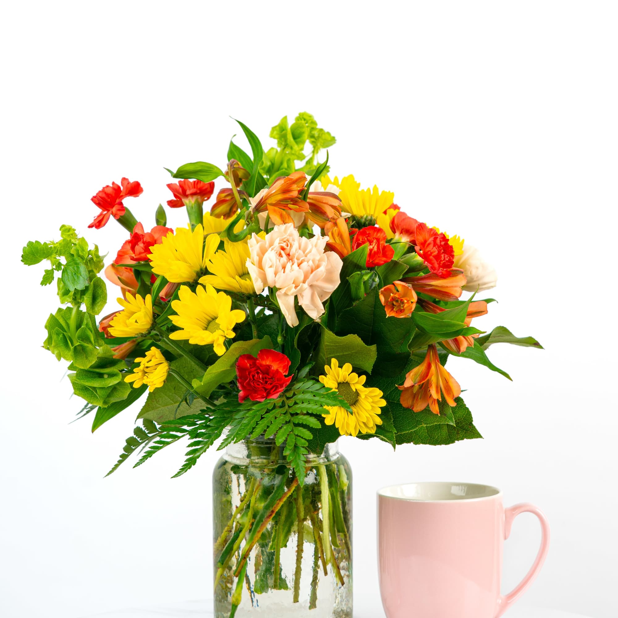 Bright mixed bouquet of yellow, orange, and red flowers in a glass jar next to a pink mug