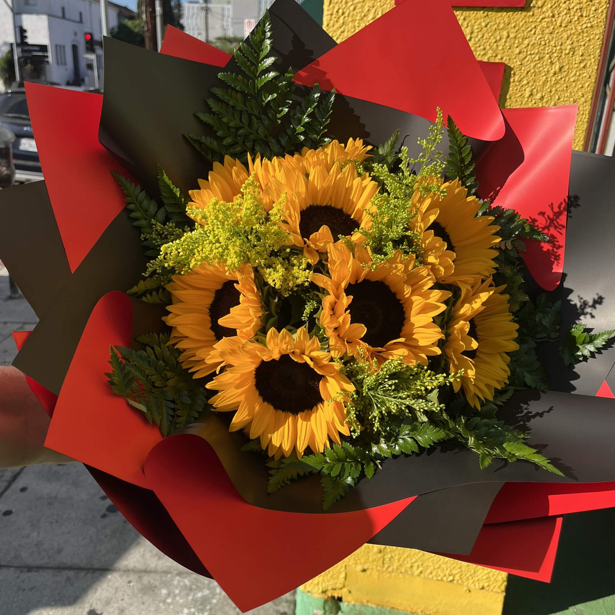 Hand-tied bouquet of yellow sunflowers with filler flowers wrapped in red and black paper.