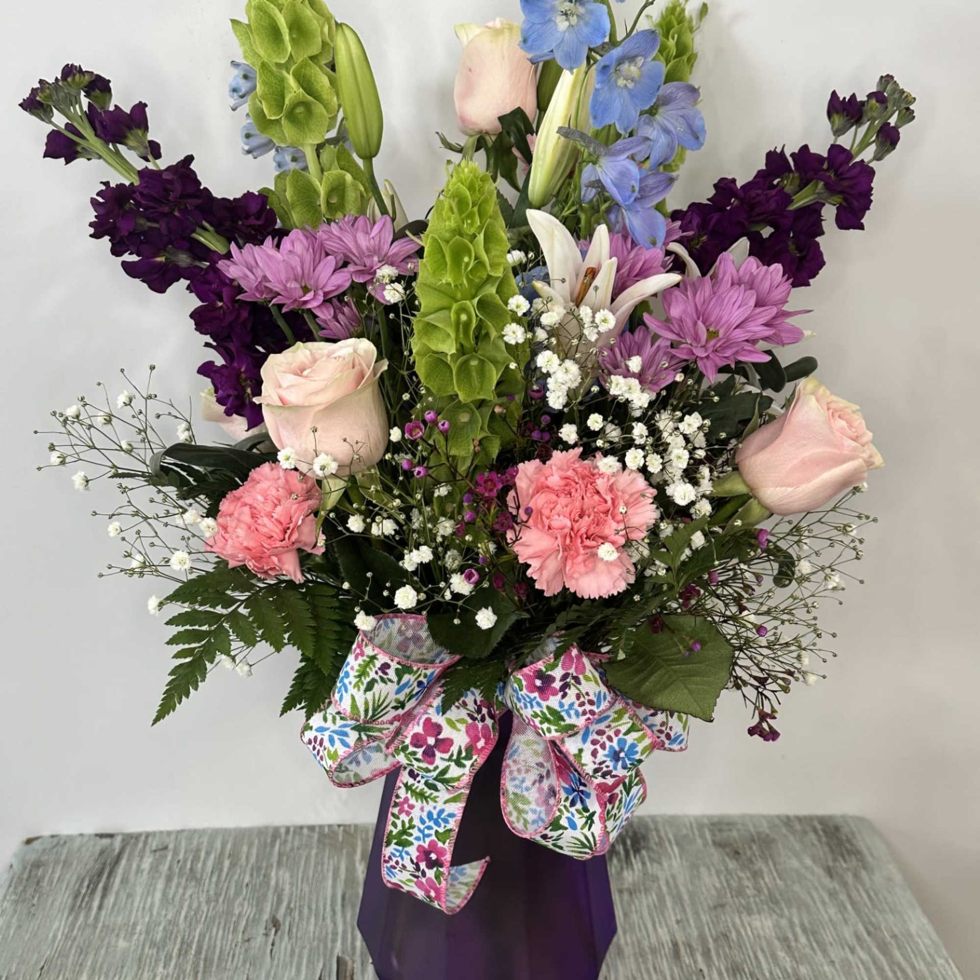 Tall mixed bouquet with pink roses, purple blooms, and blue flowers in a purple vase