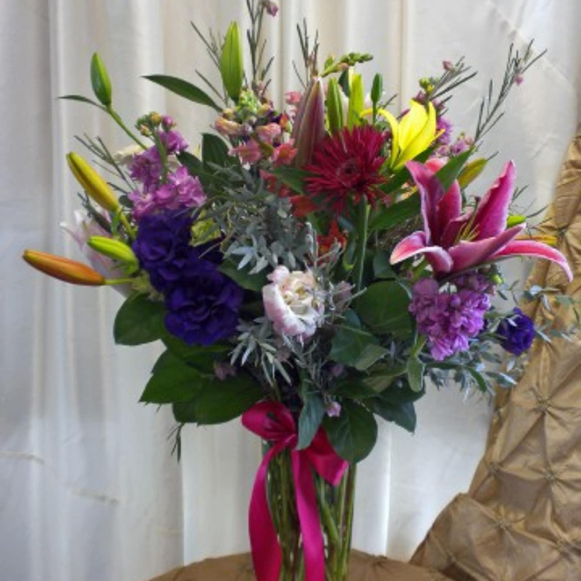 Tall mixed bouquet with lilies, purple blooms, and a pink ribbon in a glass vase