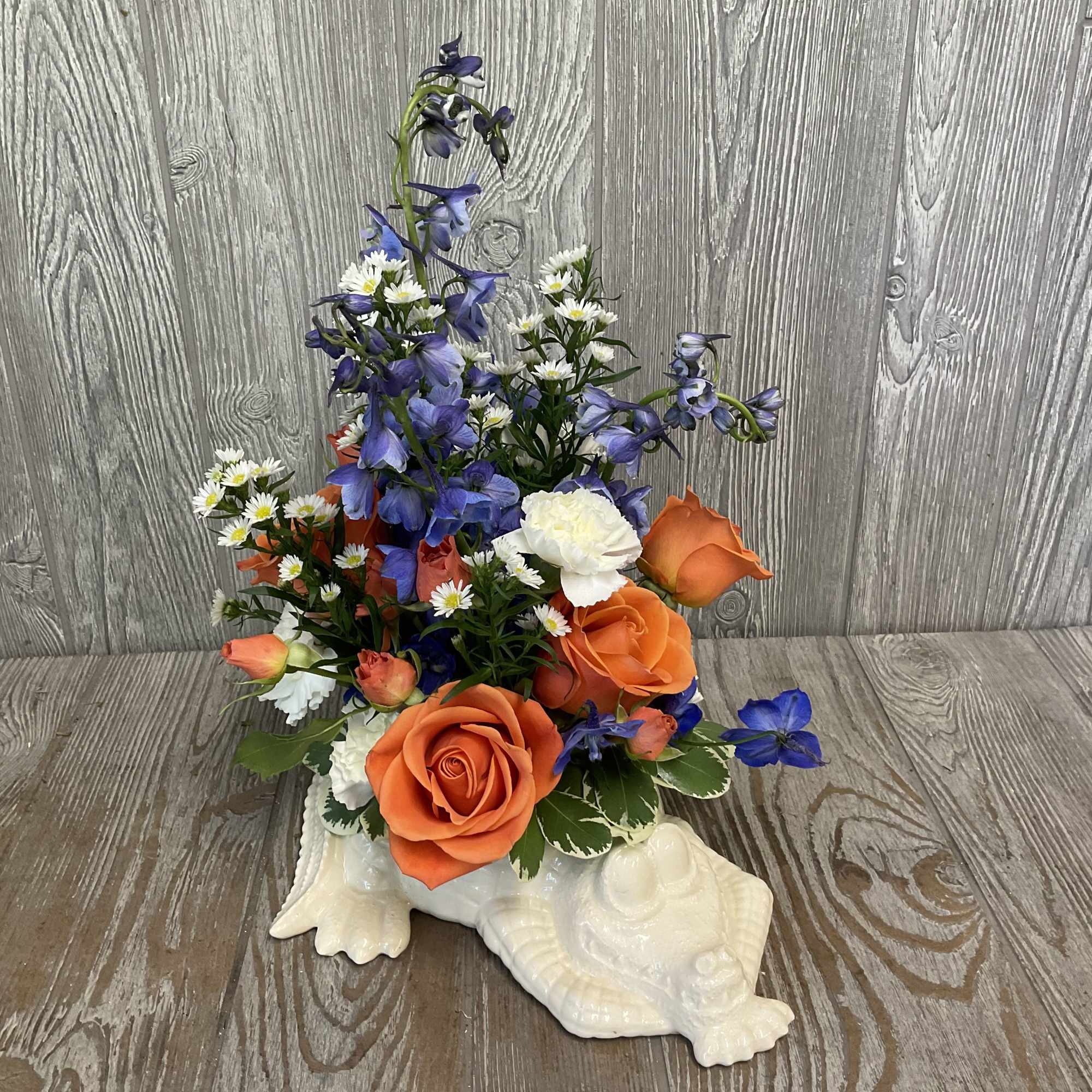 Orange roses, blue delphinium, white daisies and carnations in a white ceramic turtle-shaped container
