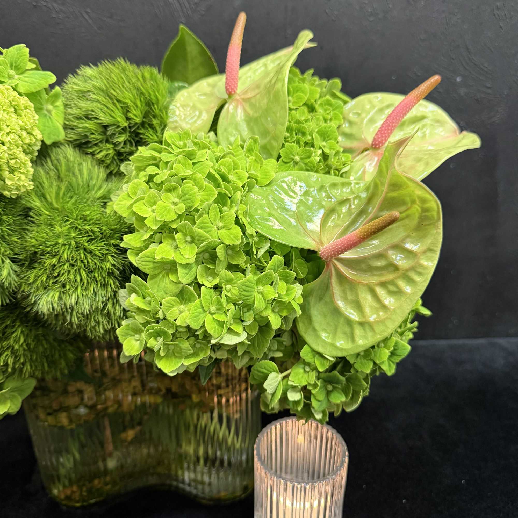Green floral arrangement with anthuriums in a glass vase beside a small candle holder