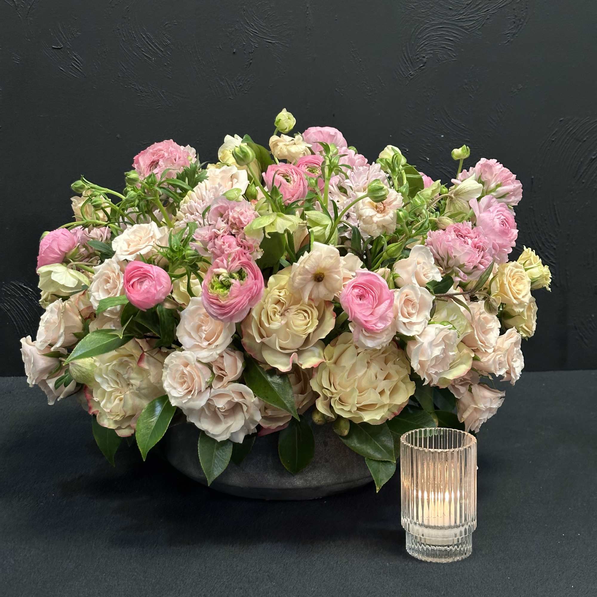 Low floral arrangement of pink and cream blooms in a dark bowl with a glass candle holder beside it
