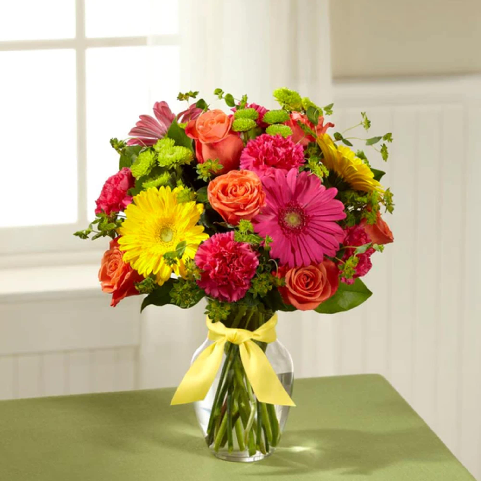 Colorful bouquet of roses, gerbera daisies, and carnations in a glass vase with a yellow ribbon
