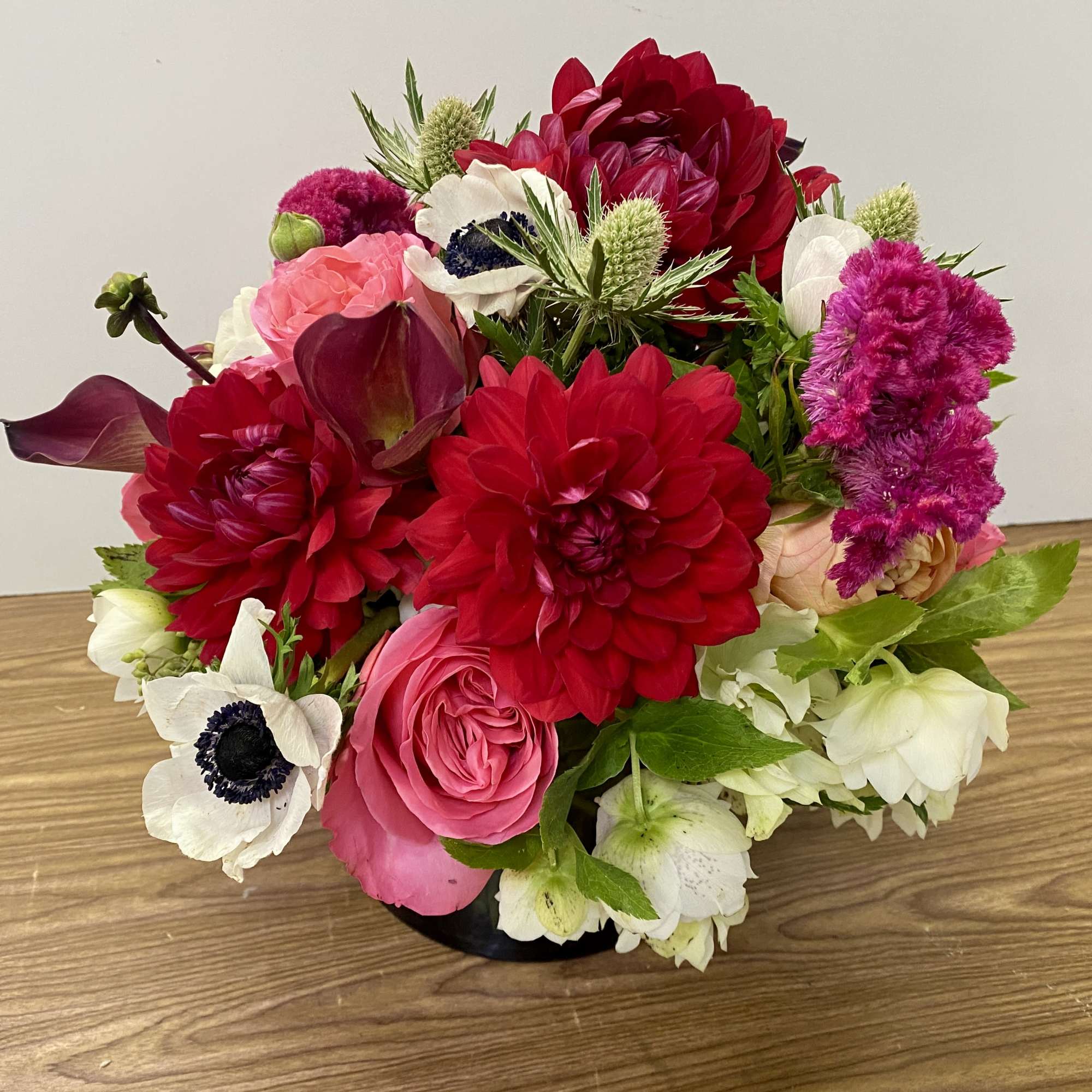 Low round arrangement of red dahlias, pink roses, white anemones and mixed blooms in a black container