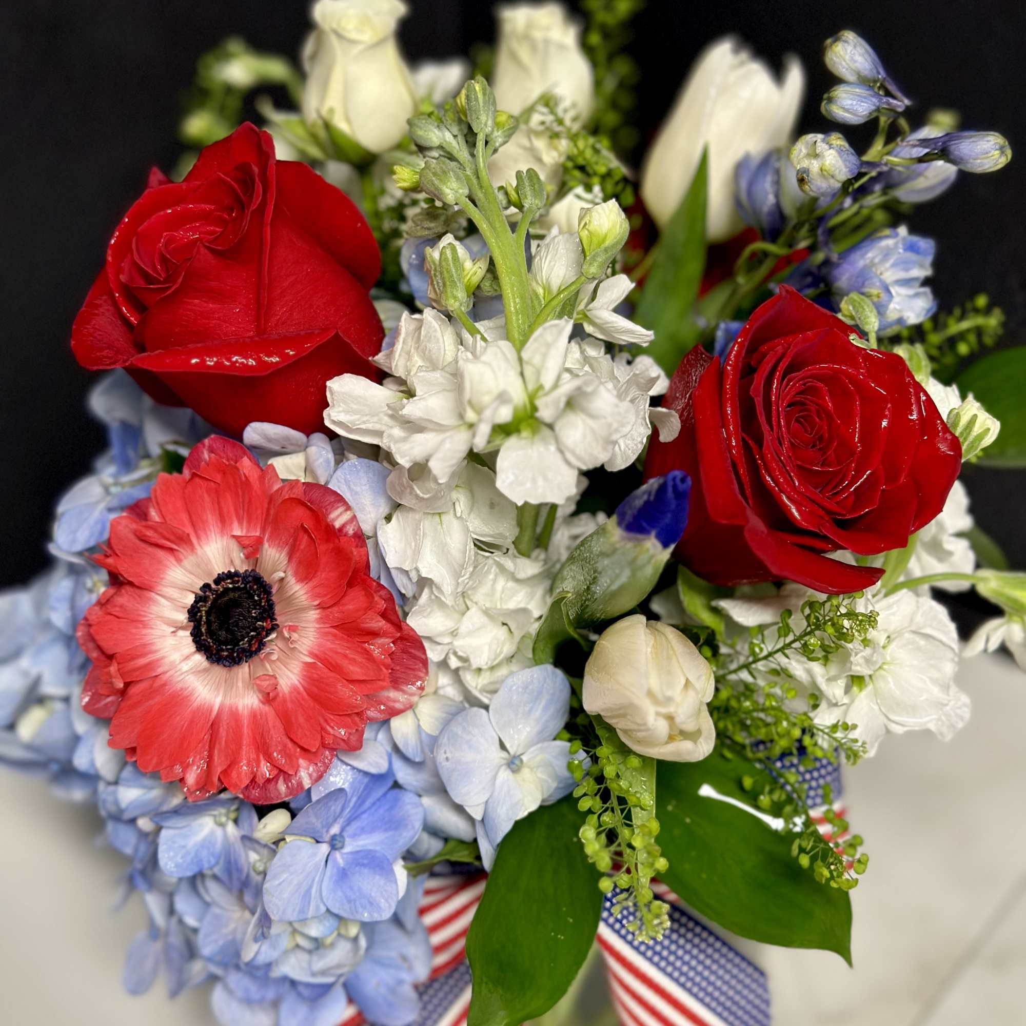 Red roses, blue hydrangeas, and white flowers in a vase with striped ribbon