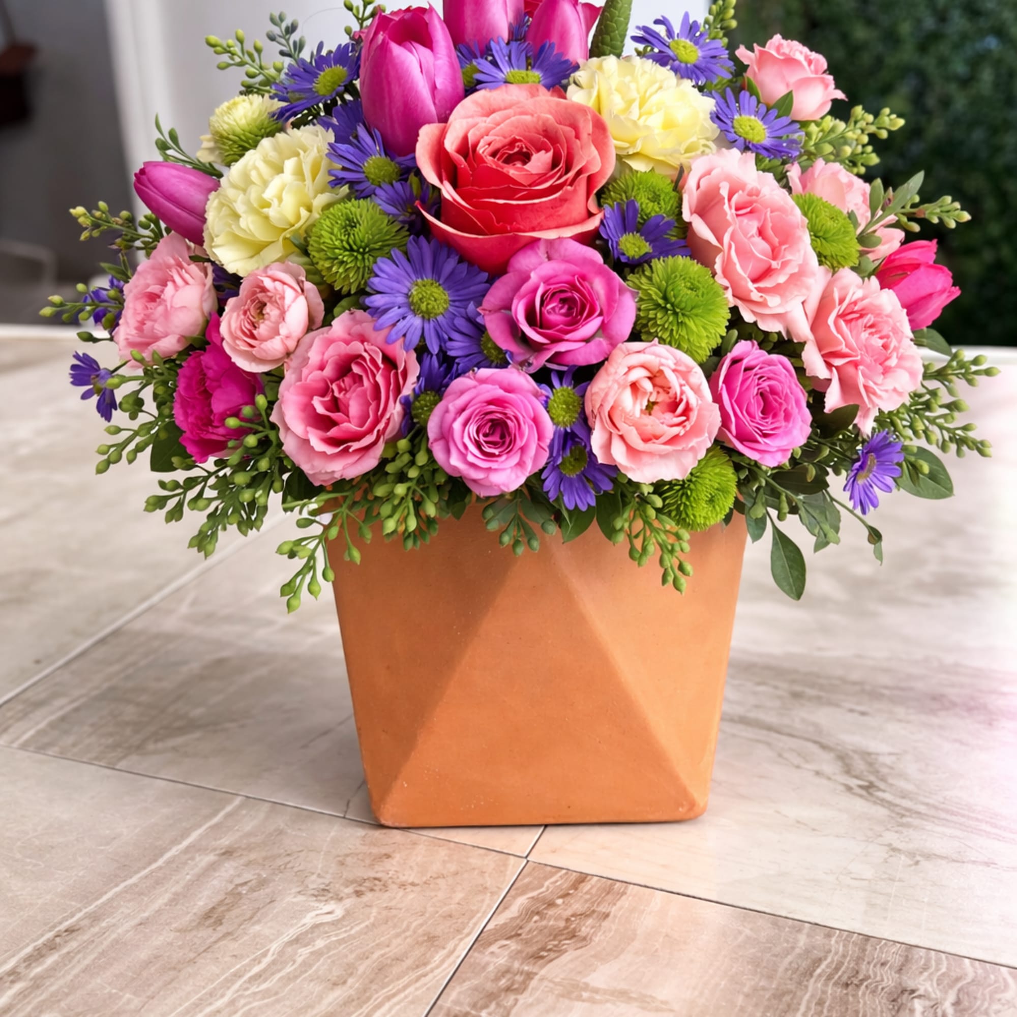 Bright mixed arrangement of pink roses, tulips and purple daisies in a terracotta cube vase
