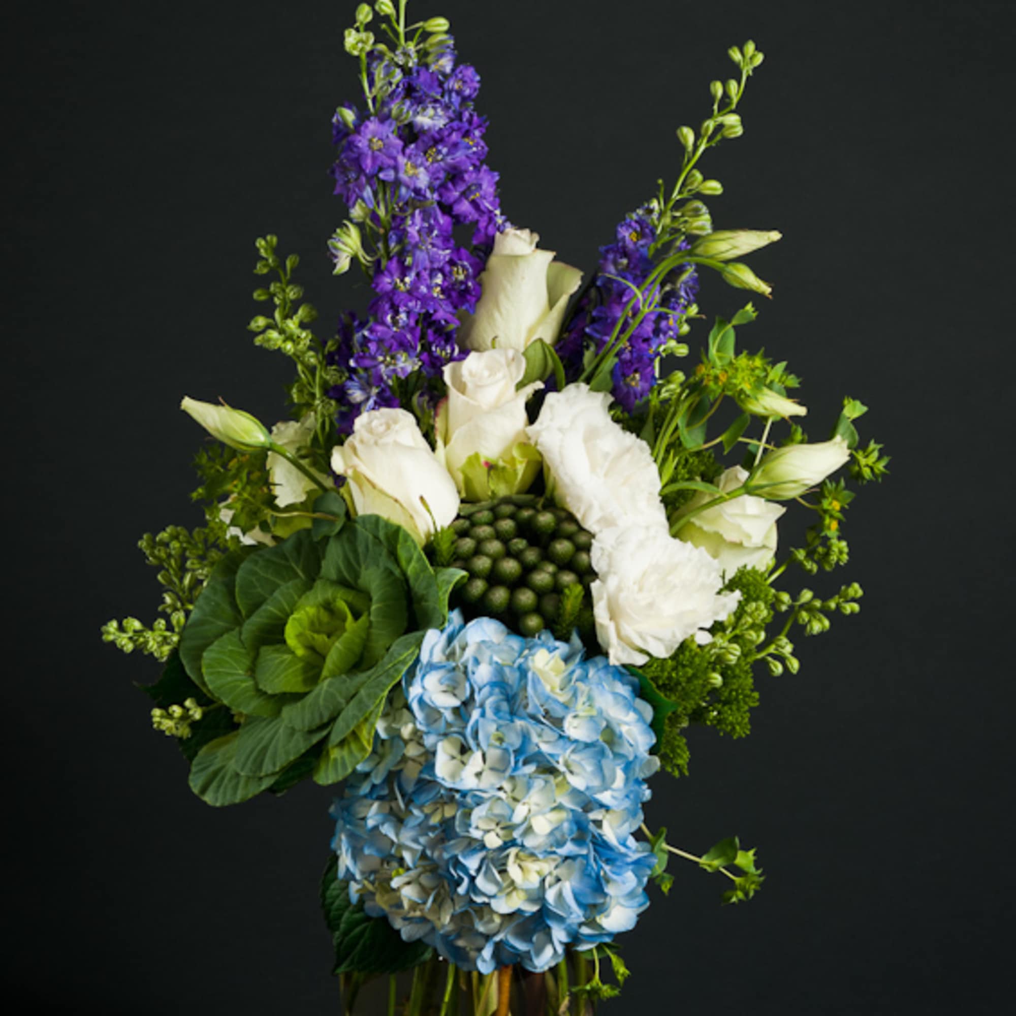 Tall bouquet of blue hydrangea, white roses, and purple flowers in a glass vase