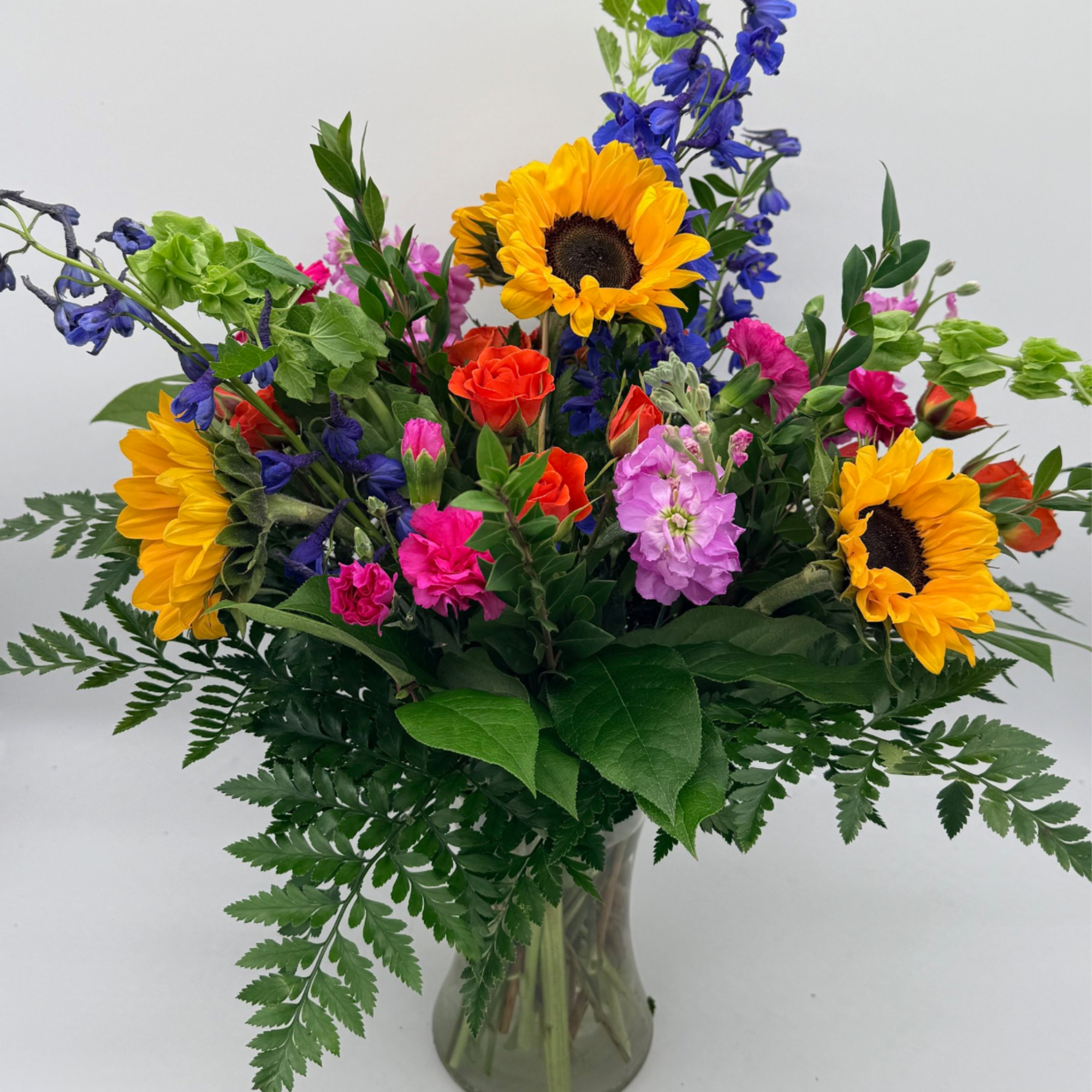 Vibrant mixed bouquet with sunflowers, pink and orange blooms, and blue delphinium in a clear glass vase.