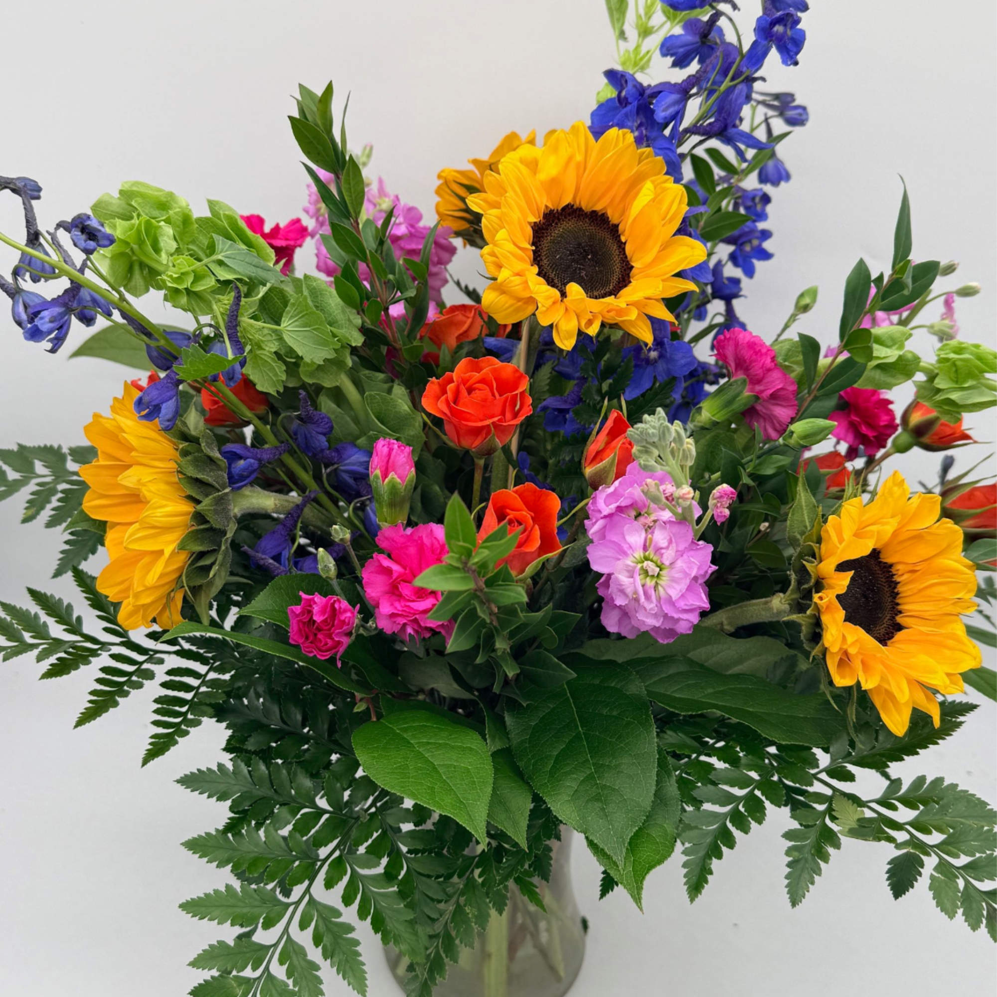 Mixed arrangement of bright sunflowers, pink and orange blooms, and blue delphinium in a glass vase