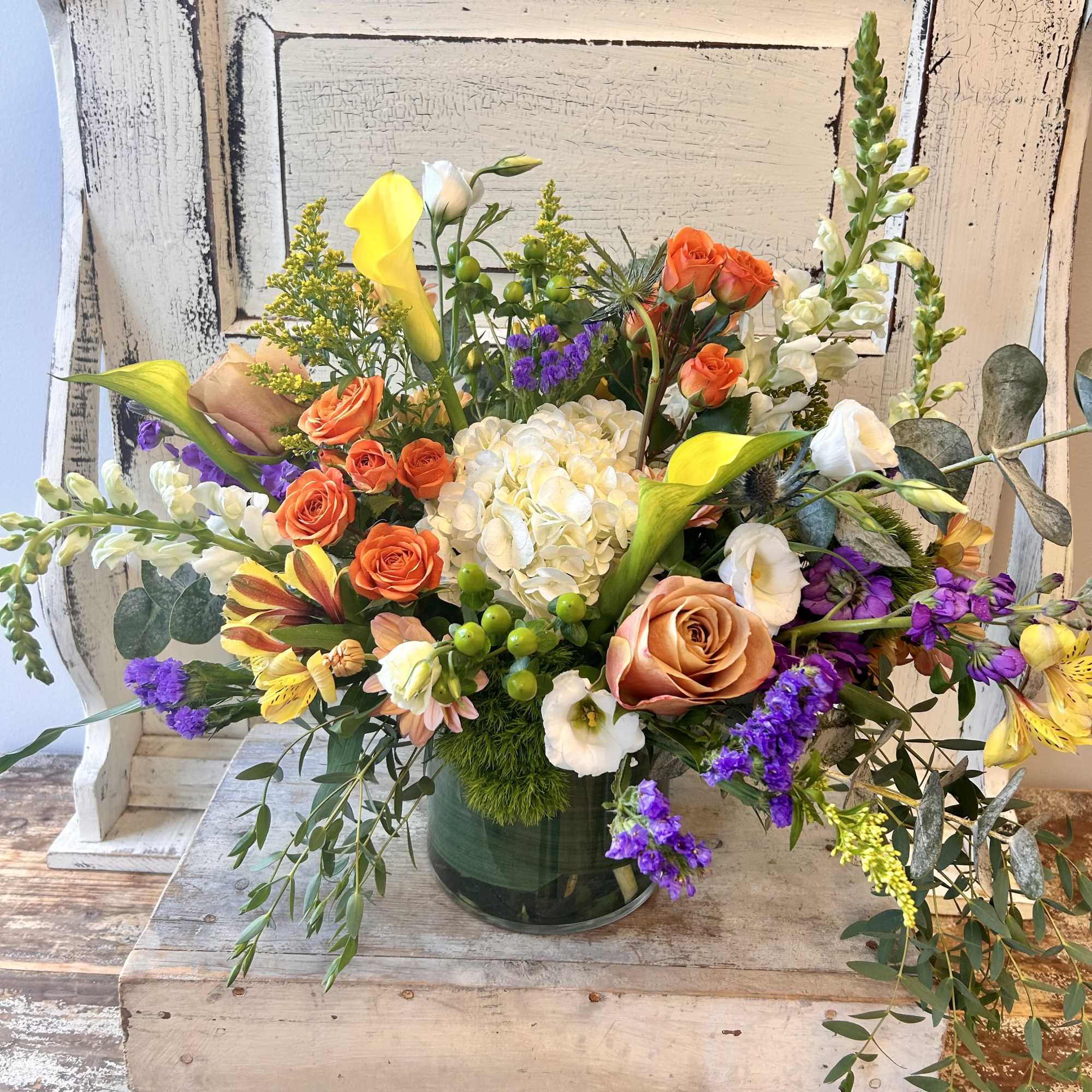 Mixed bouquet in a glass vase with orange roses, yellow calla lilies, and purple accents