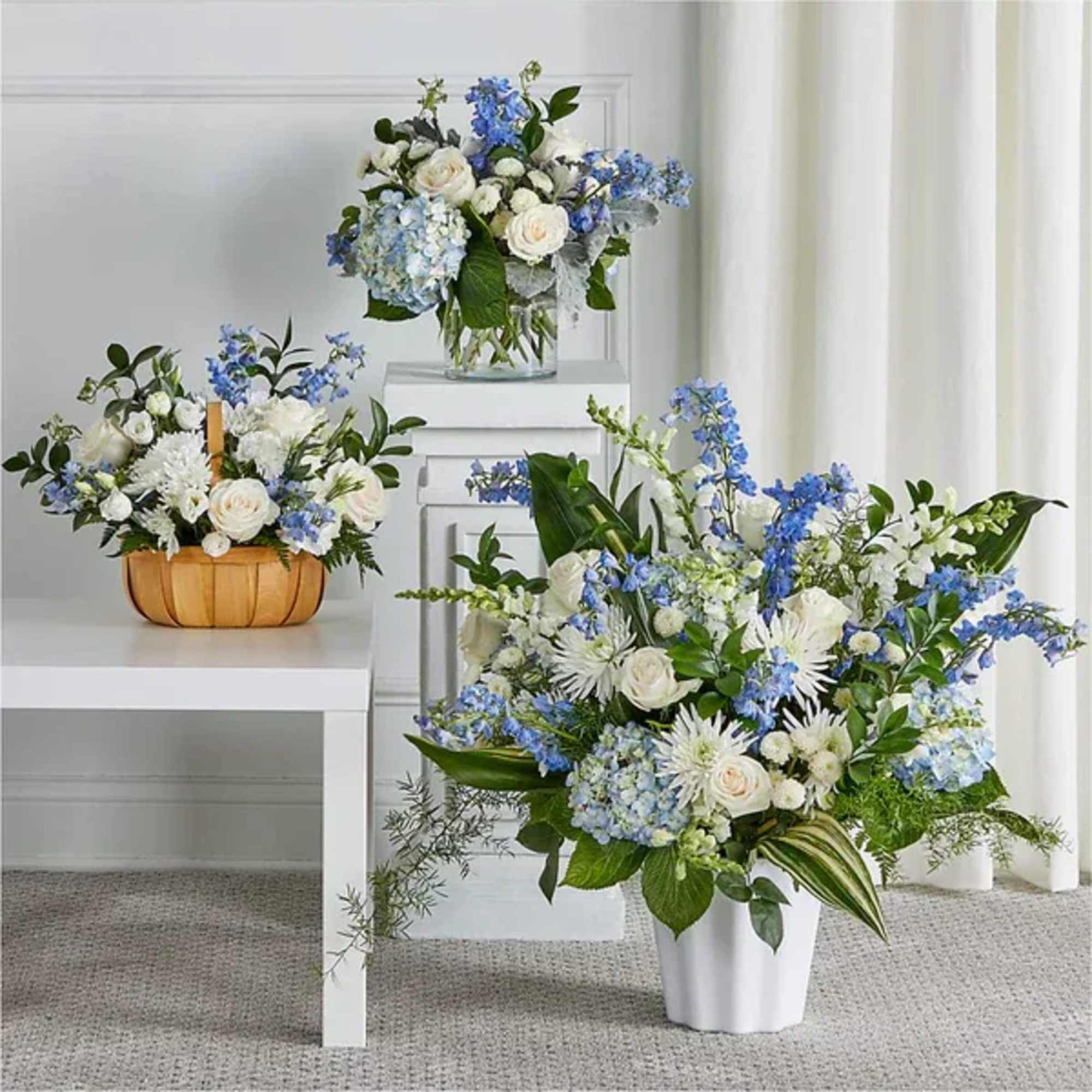 Set of three blue and white flower arrangements in basket, glass vase, and white urn.