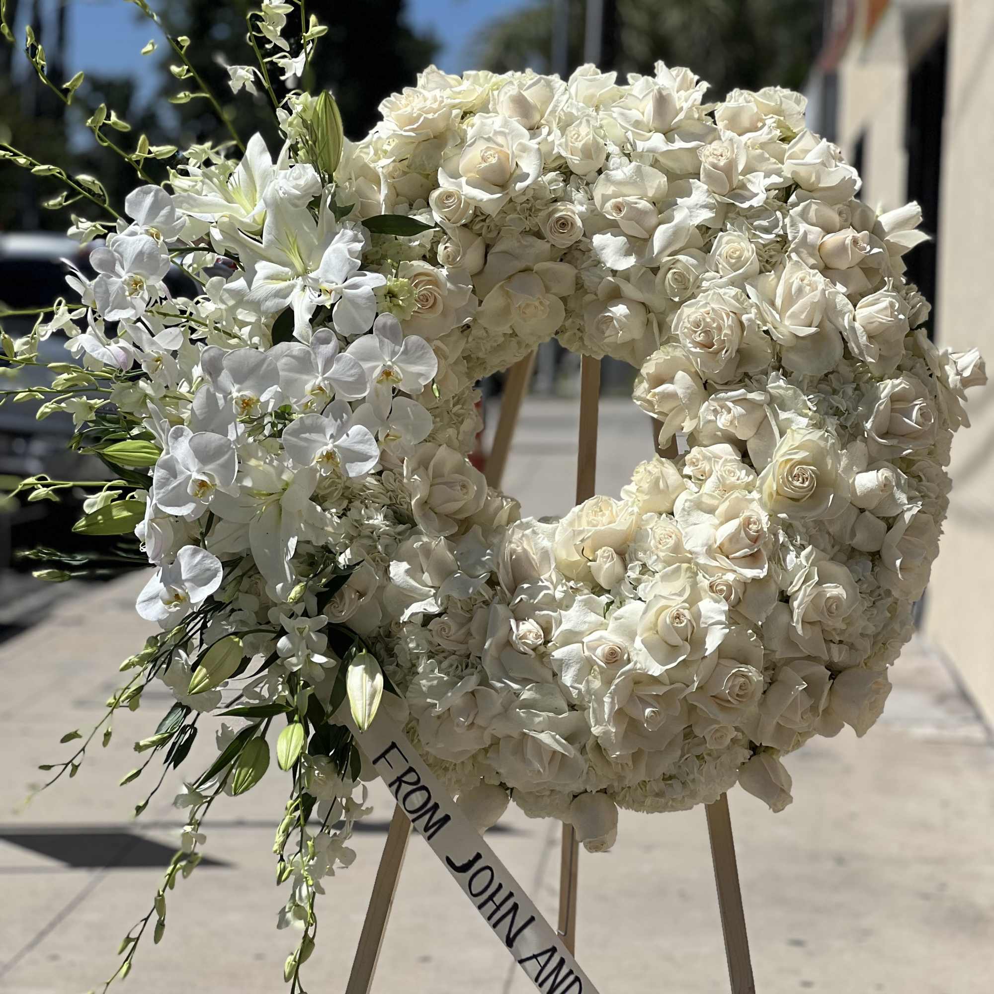 White floral wreath on an easel with a ribbon reading "FROM JOHN AND NICOLE"