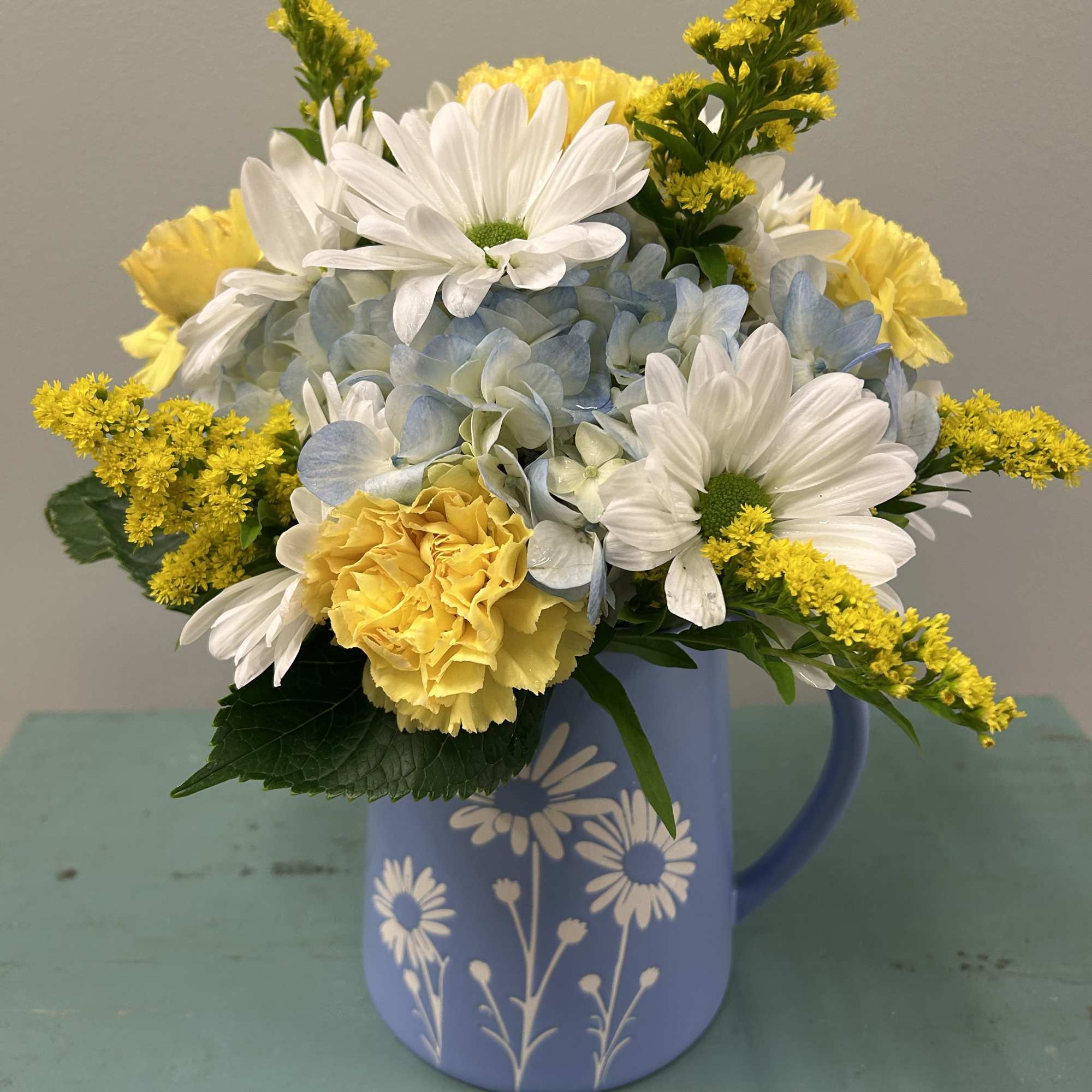 Bright, cheerful daisies and sunny yellow carnations mingle with soft blue hydrangea