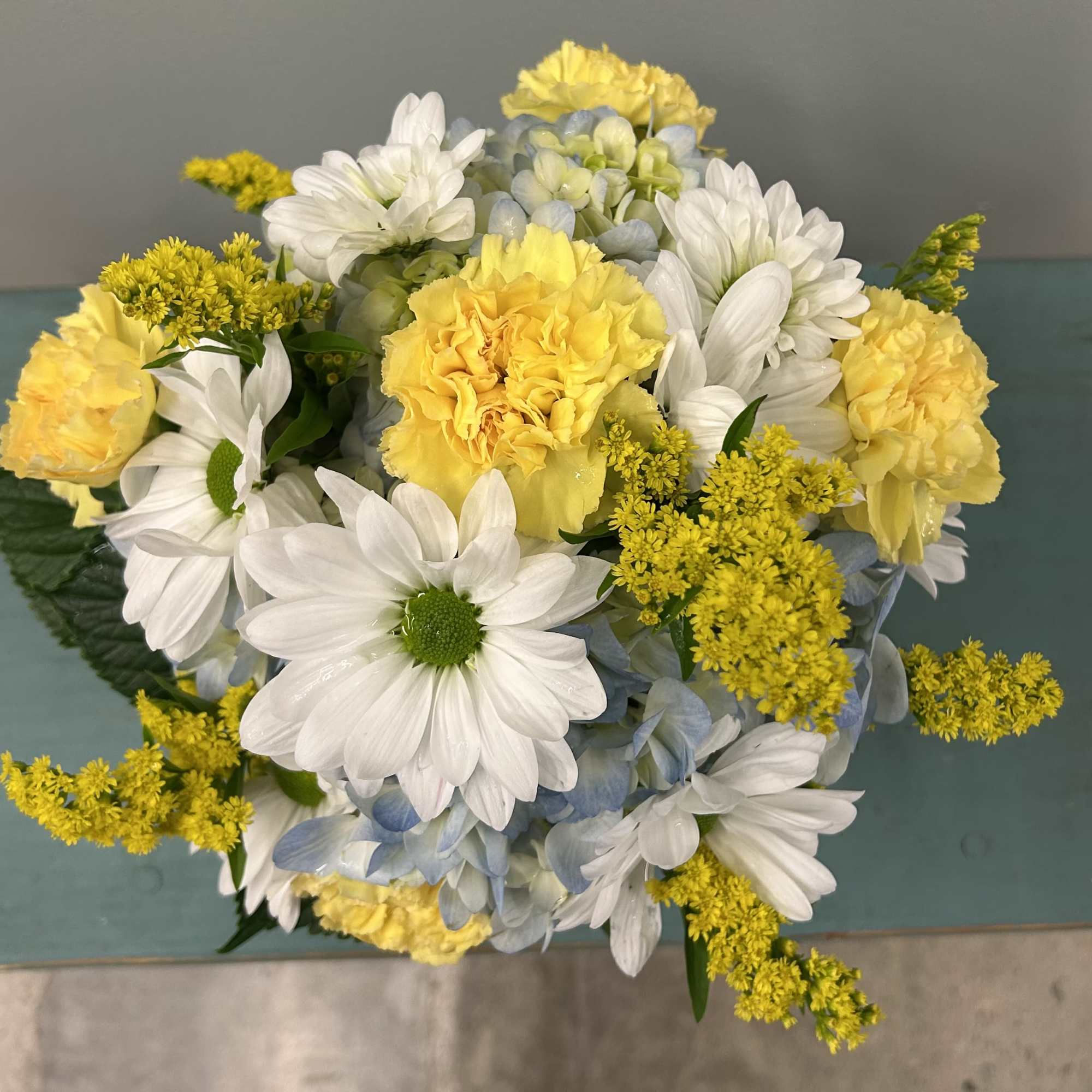 Bright, cheerful daisies and sunny yellow carnations mingle with soft blue hydrangea