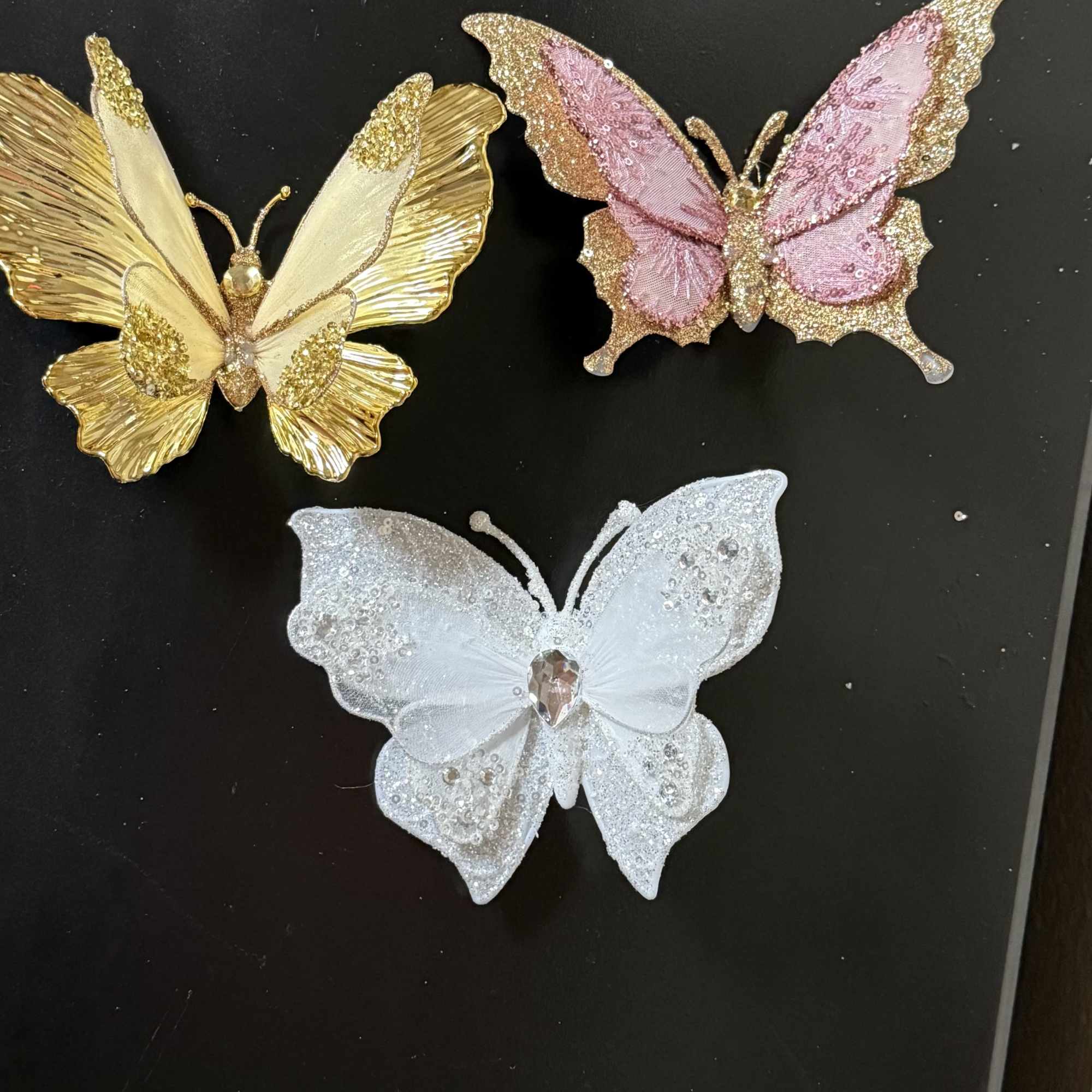 Three glittery butterfly ornaments in gold, pink, and white on a black surface