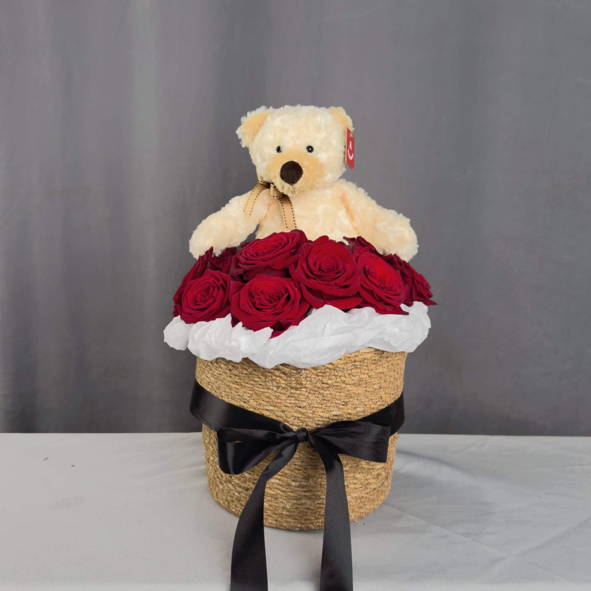Teddy bear sitting in a basket of red roses with a black ribbon