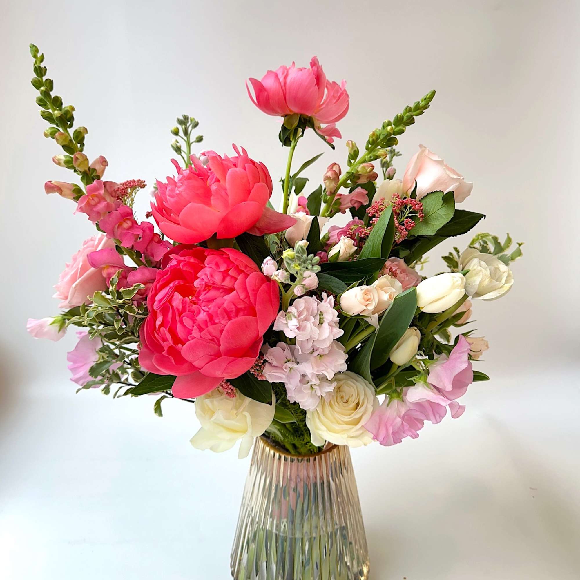 Vase arrangement of bright pink peonies with pale roses, stock, and snapdragons in a ribbed glass vase