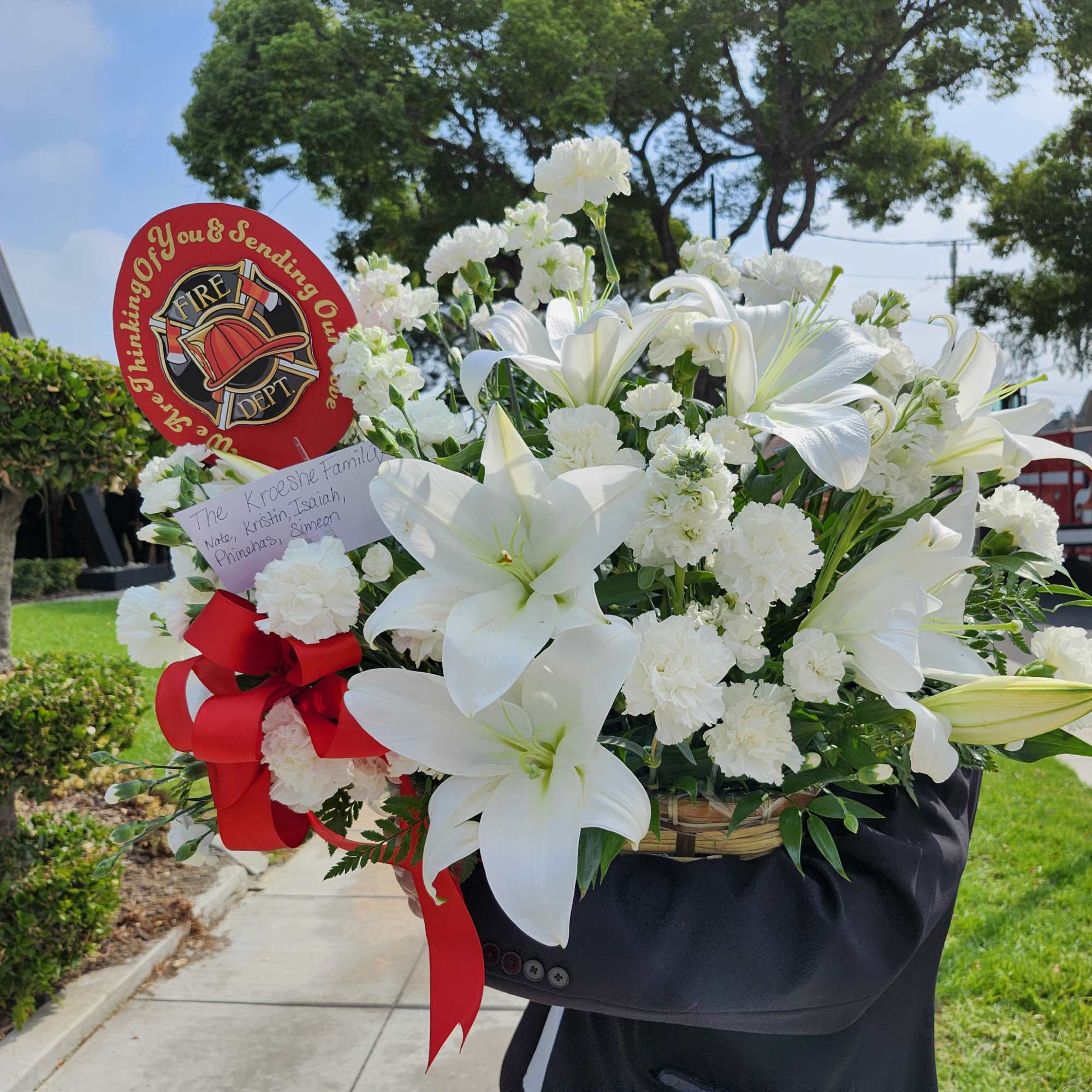 White lilies and carnations in a basket with a red fire department tribute sign.