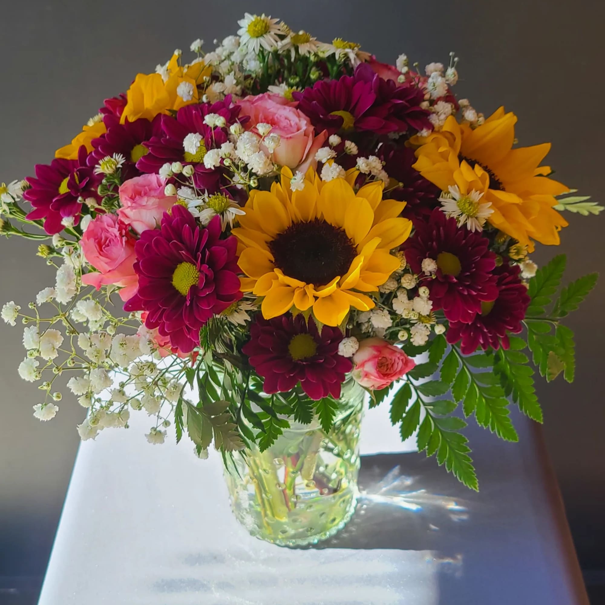 Bouquet of sunflowers, pink roses, and dark magenta daisies in a glass vase