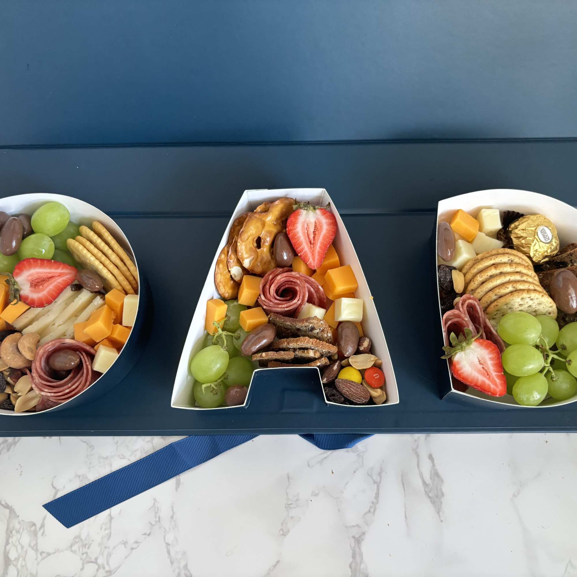 Three letter-shaped snack boxes filled with fruit, cheese, crackers, and meats