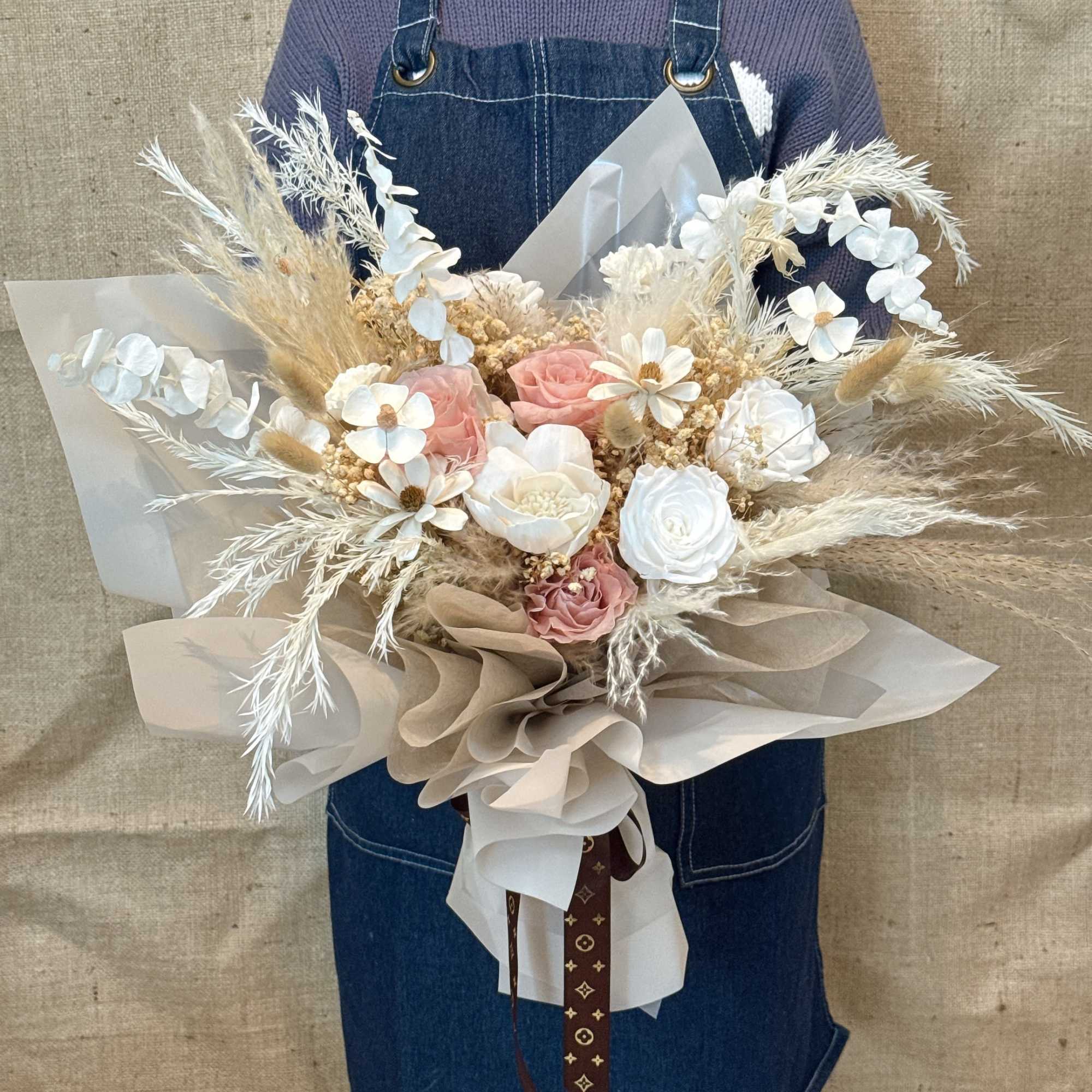 Large bouquet of soft pink and white roses with dried grasses wrapped in layered beige paper.