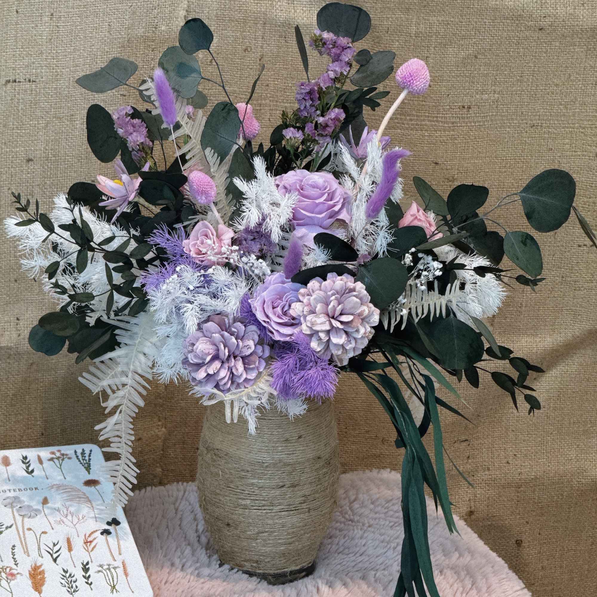Lavender and pink flower arrangement with dark foliage in a twine-wrapped vase on a soft pink surface