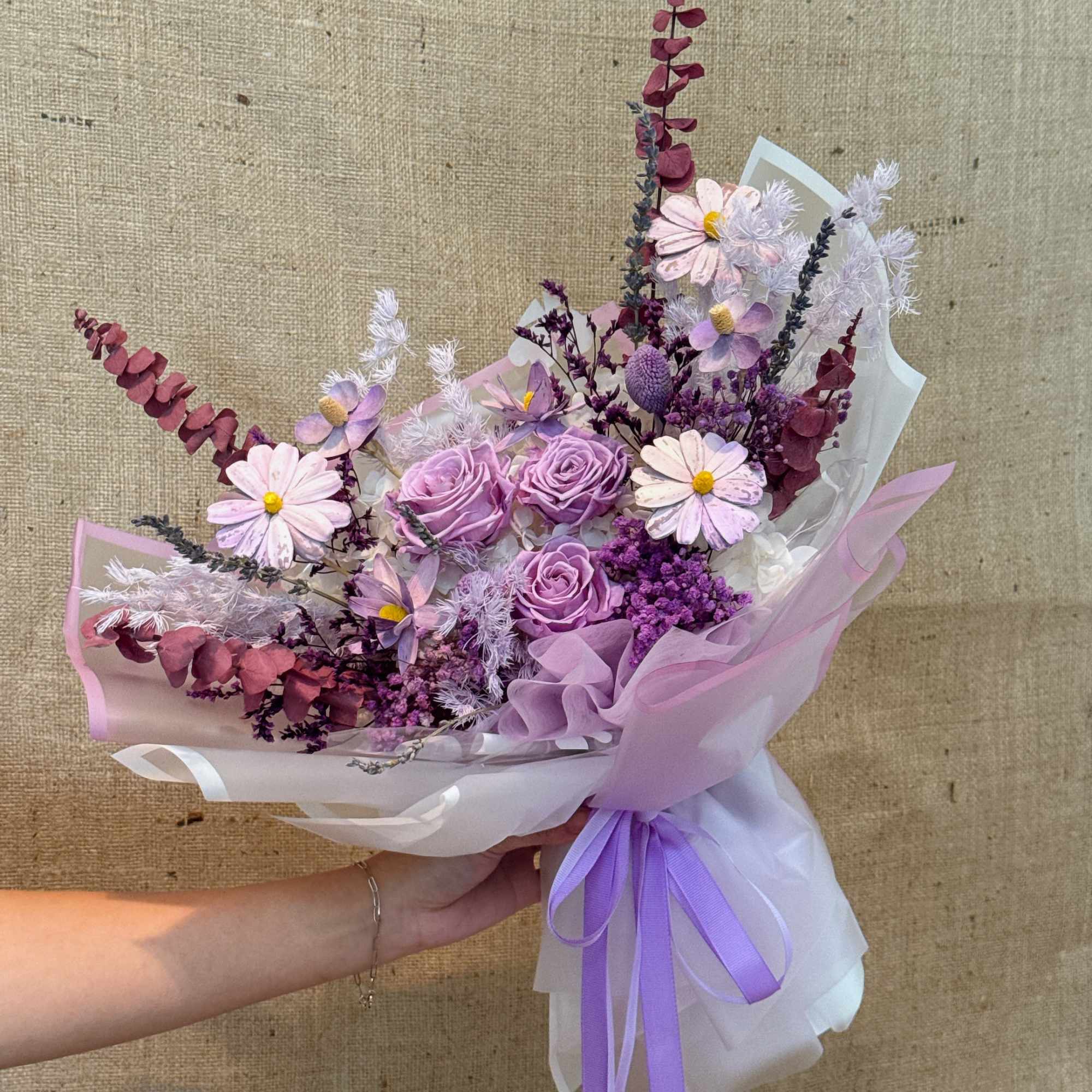Hand-tied bouquet of lavender roses and pink daisy-like flowers wrapped in white and lilac paper with purple ribbons.