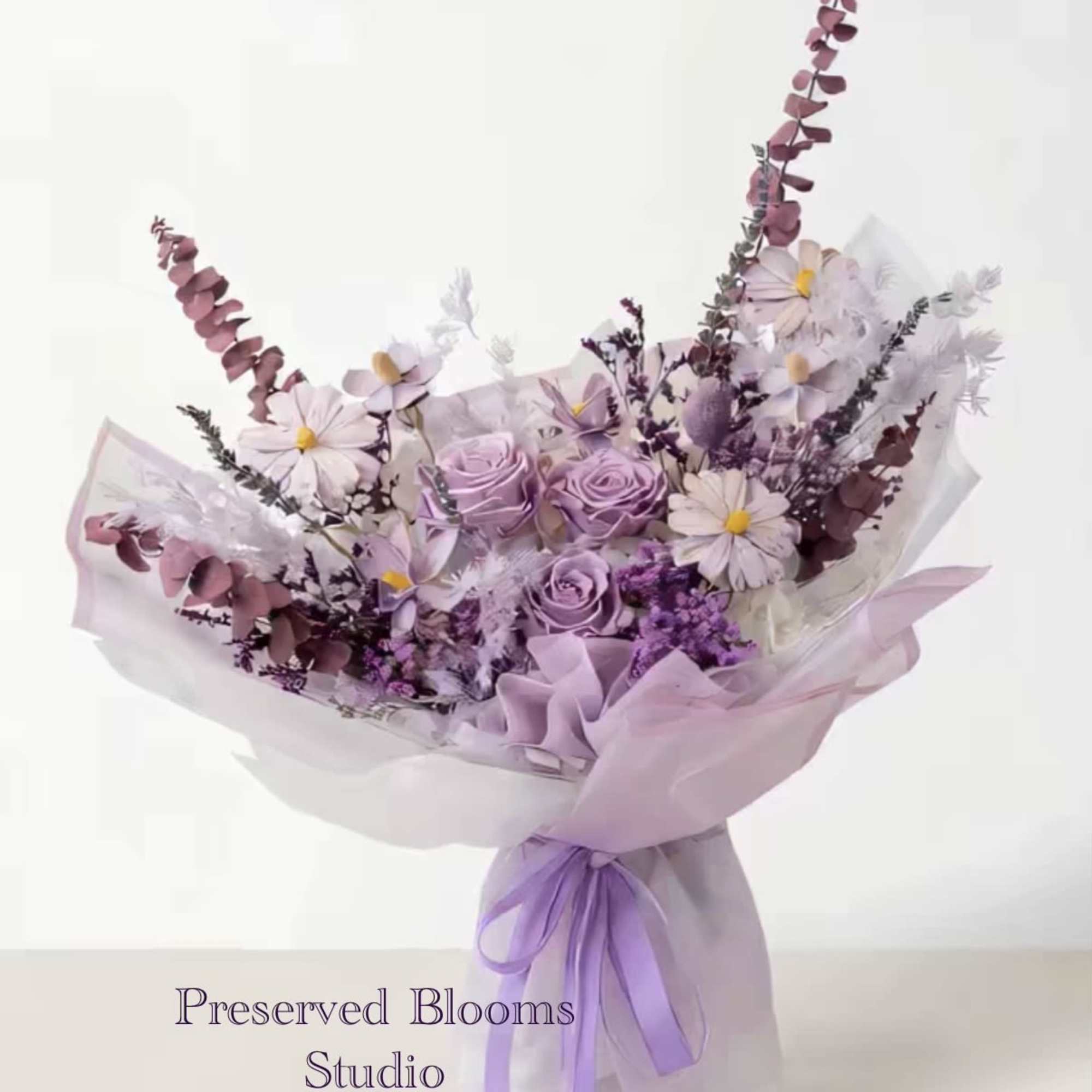 Lavender and white bouquet of roses and daisies wrapped in sheer paper with a purple ribbon bow