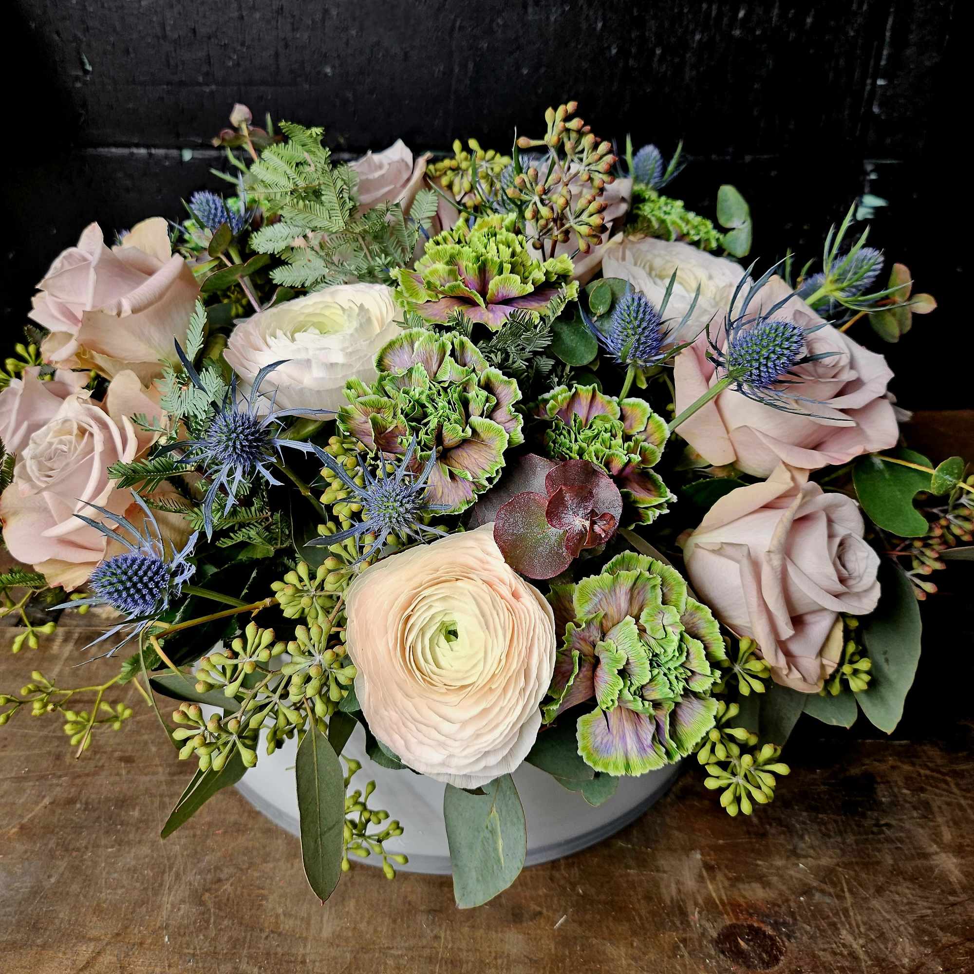 Bouquet of blush roses, white ranunculus, and blue thistle in a white container