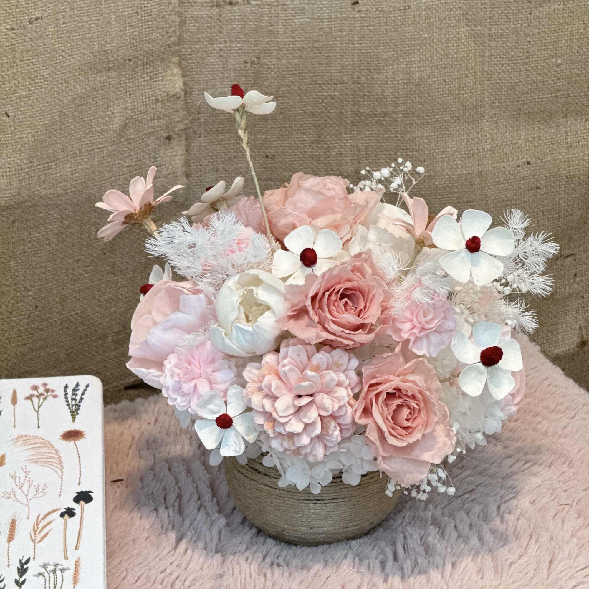 Pastel pink and white artificial flower arrangement in a small twine-wrapped pot on a soft pink surface.