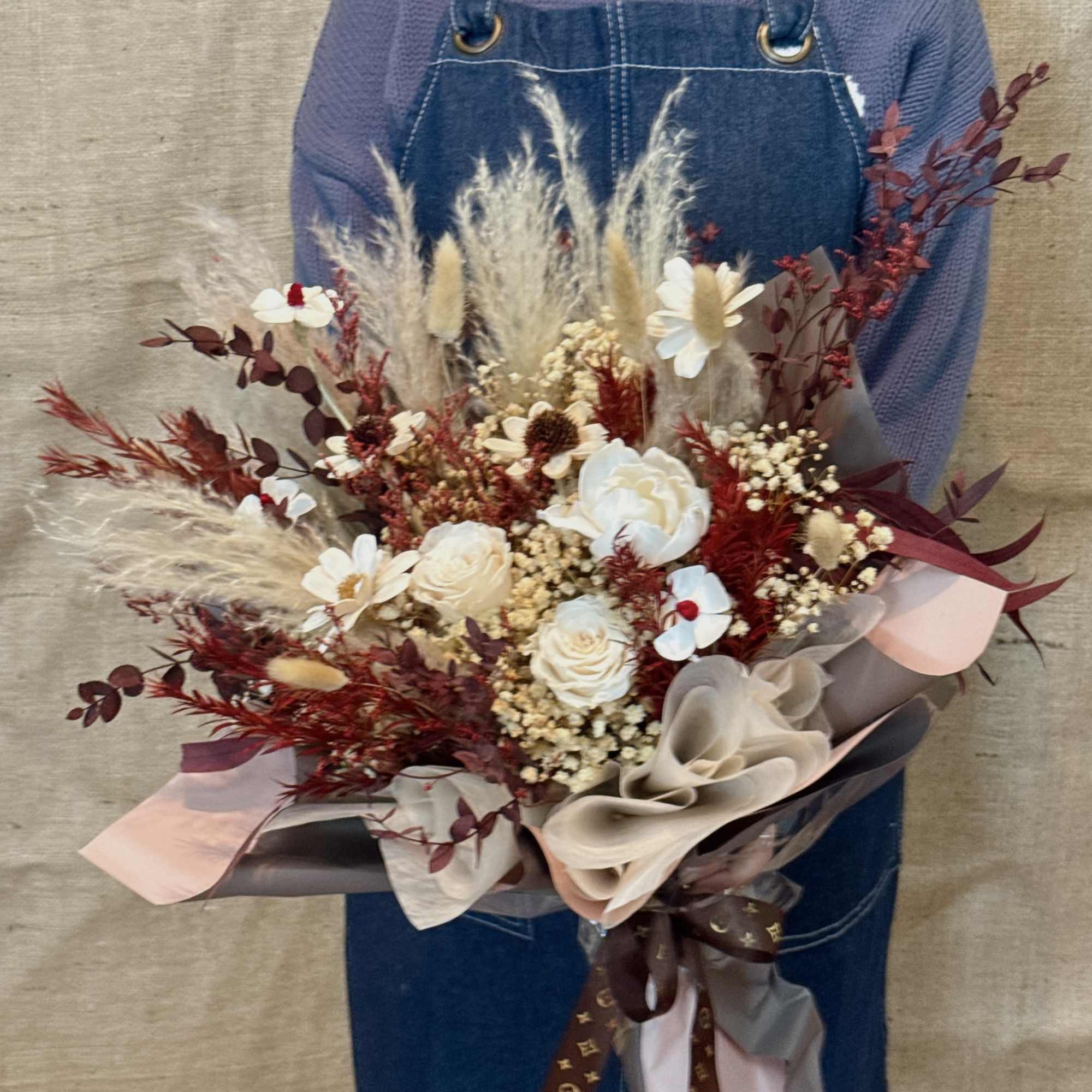 Large rustic bouquet of cream roses, dried grasses, and burgundy accents wrapped in layered paper with a bow.