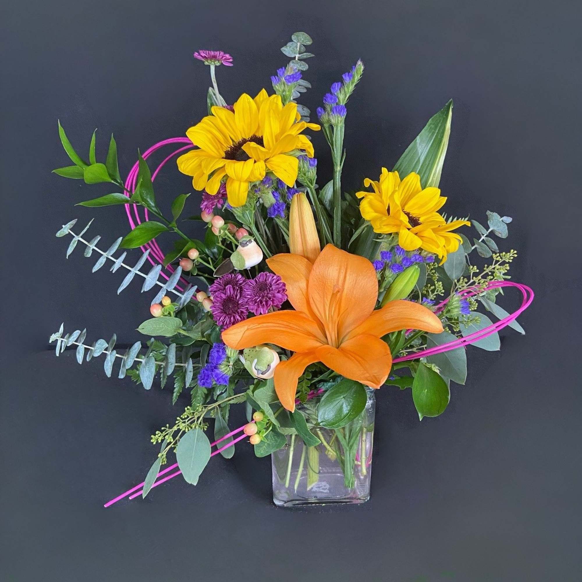 Bright mixed bouquet with sunflowers, orange lily, and purple accents in a glass vase