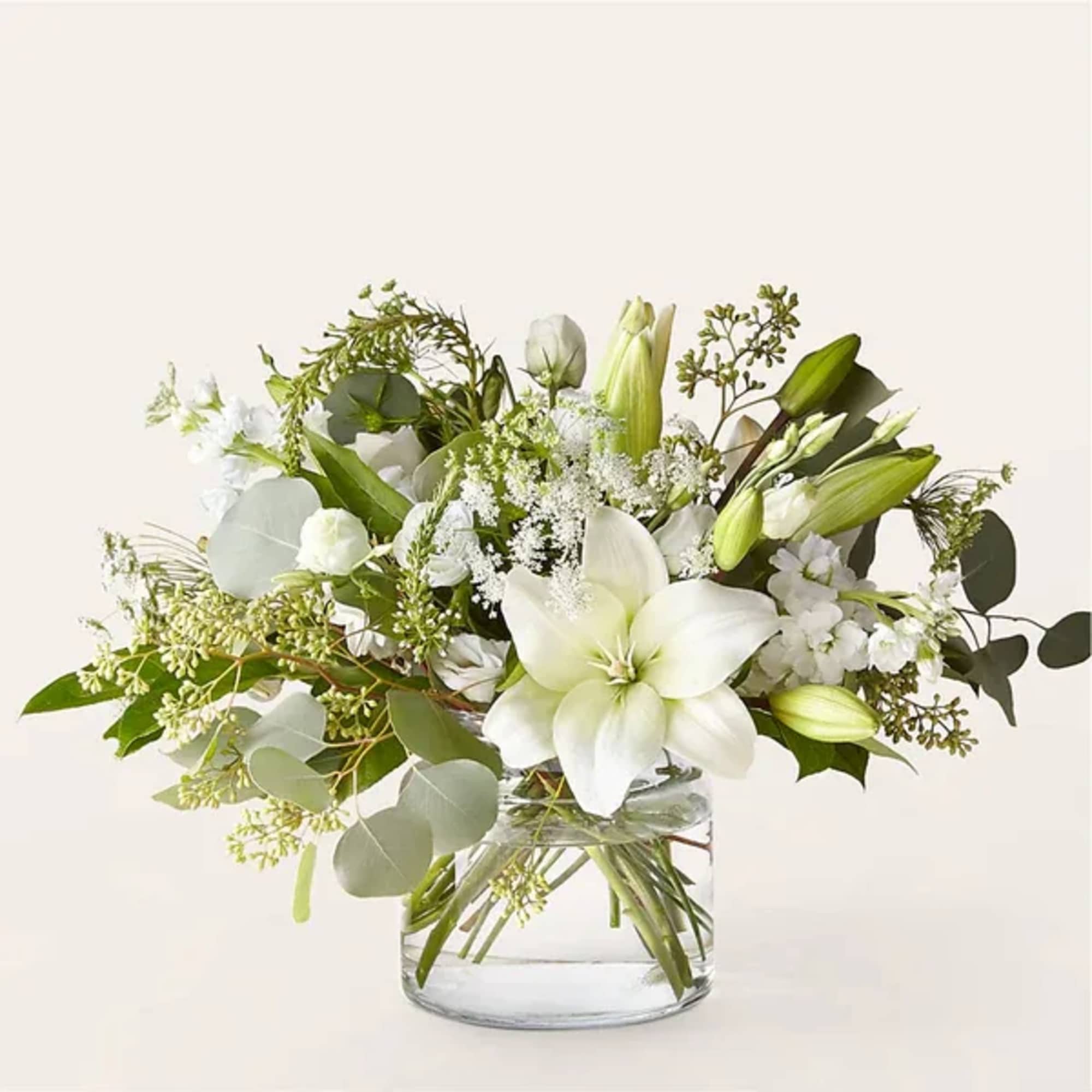 Low glass vase of white lilies and mixed white flowers with abundant foliage in a loose arrangement