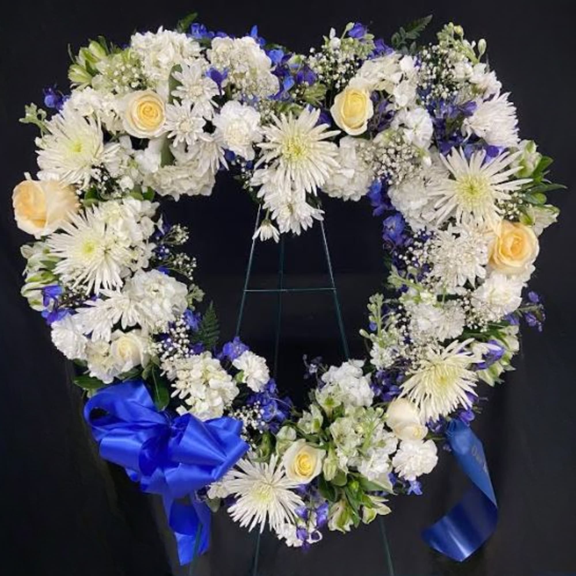 Heart-shaped wreath of white and cream flowers with blue accents on a stand