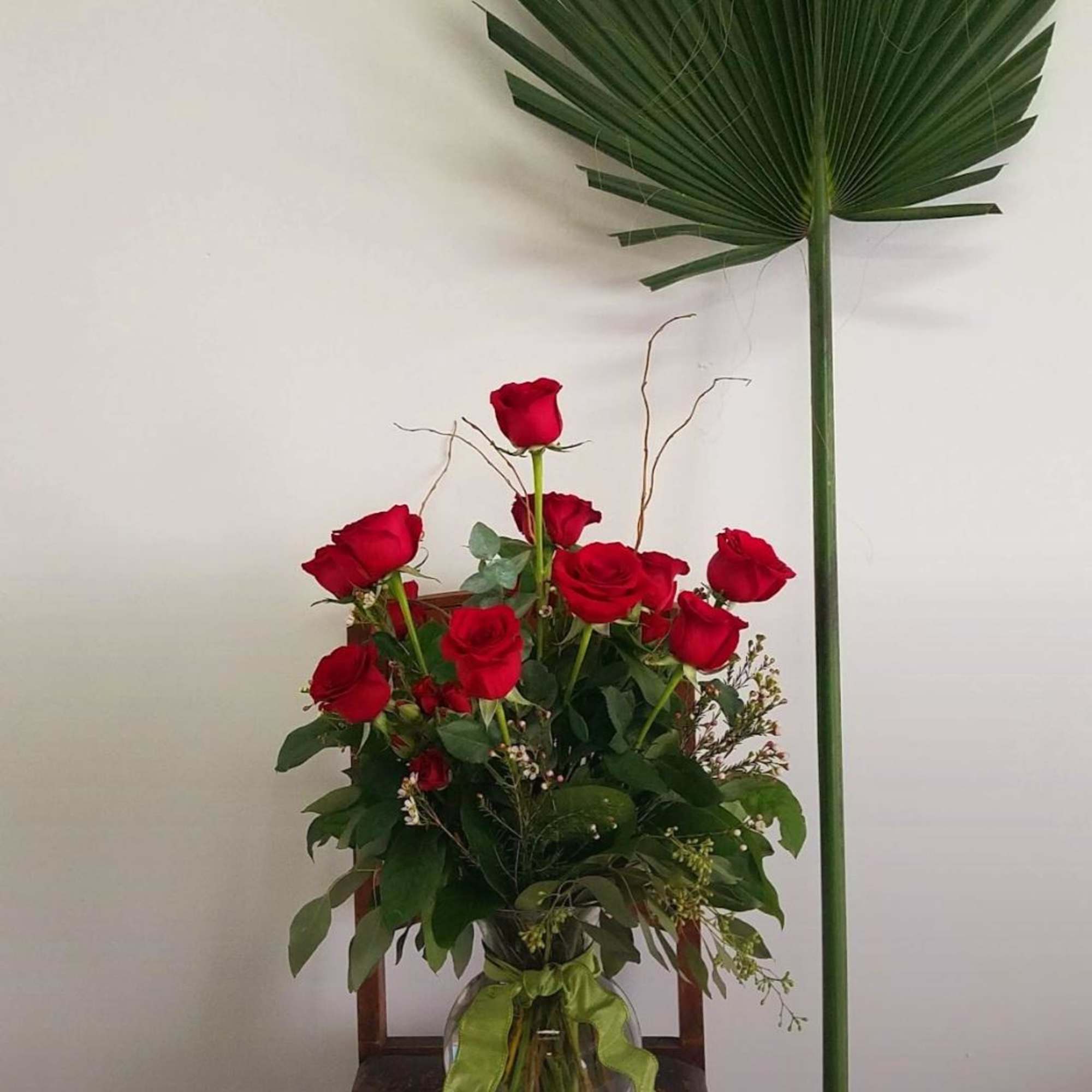 Red roses in a glass vase beside a tall palm frond.