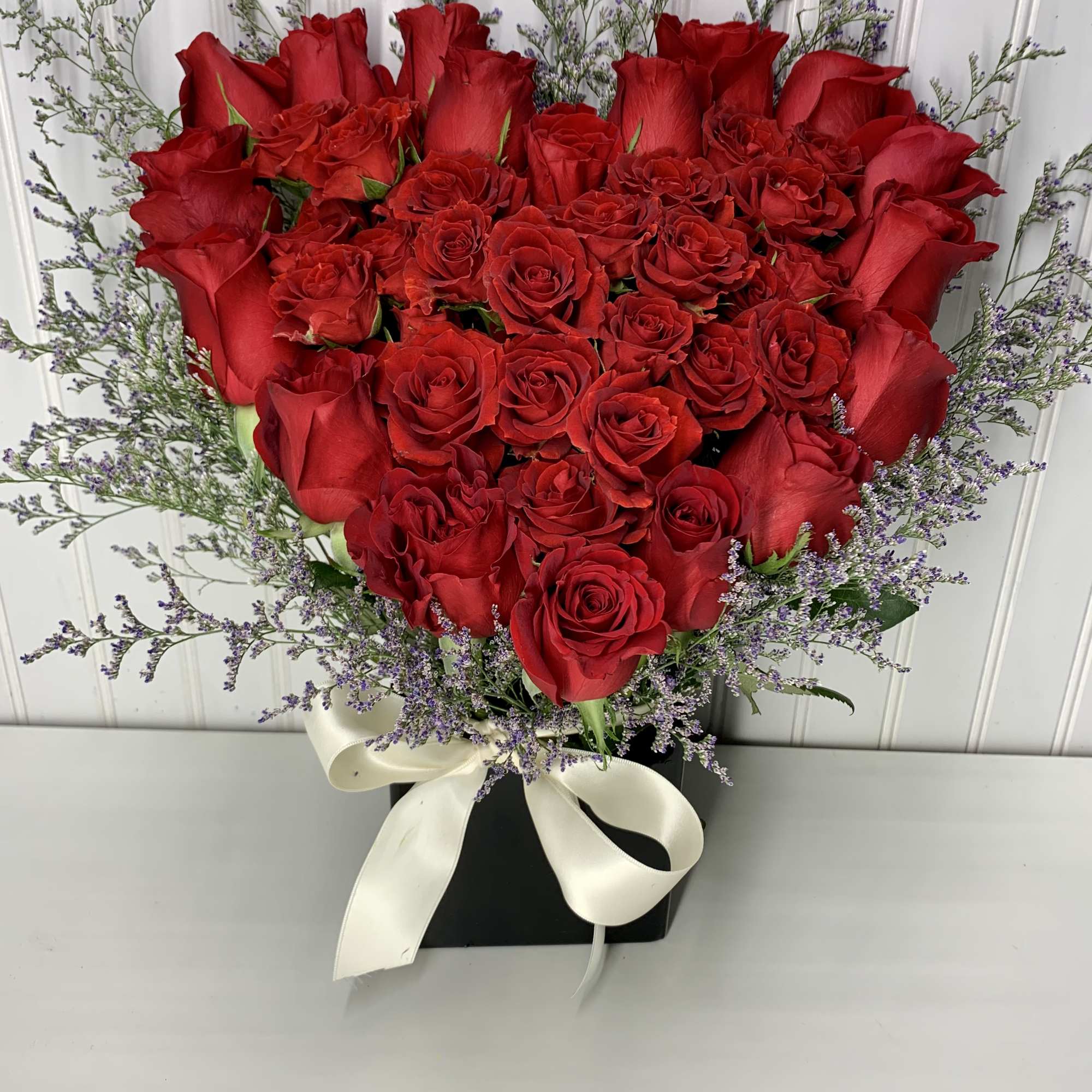 Heart-shaped arrangement of red roses in a black box with purple filler flowers and a white bow.