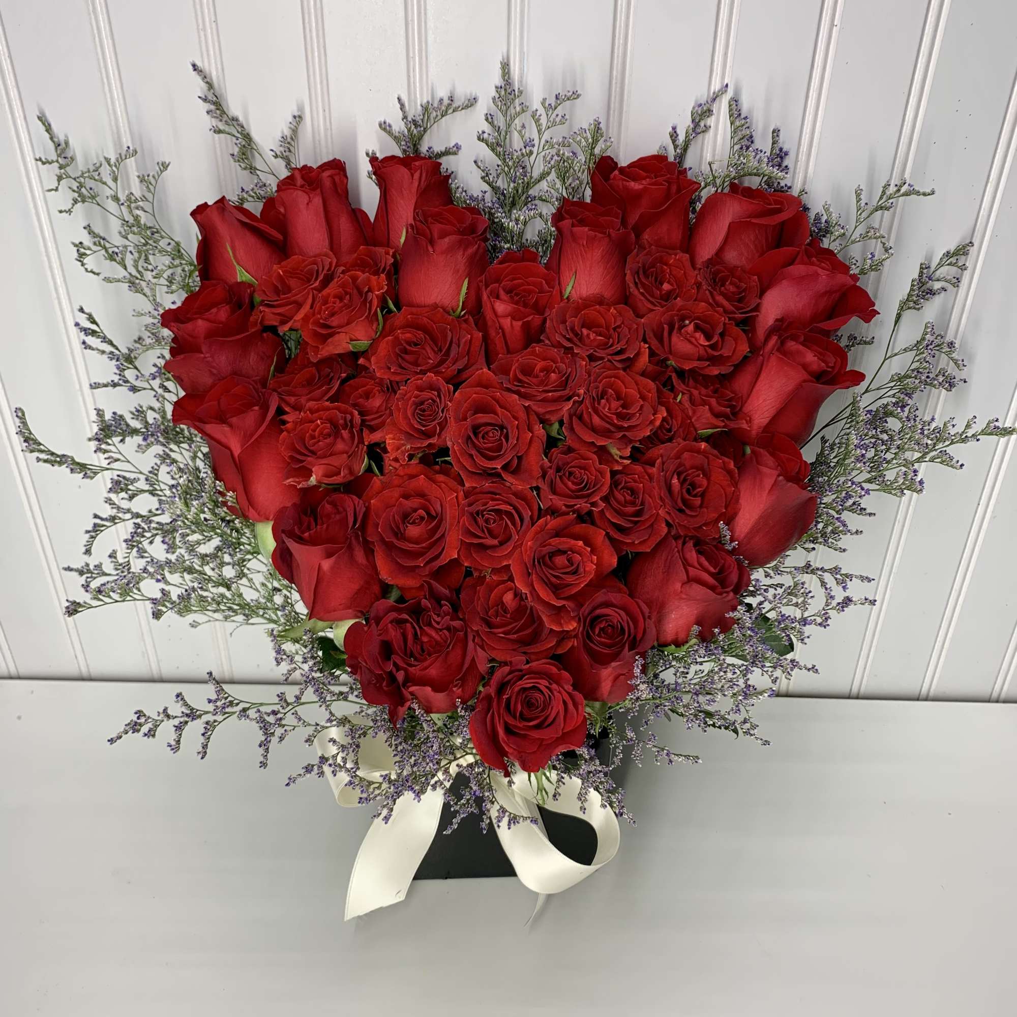 Heart-shaped arrangement of red roses with purple filler in a black box with white ribbon.